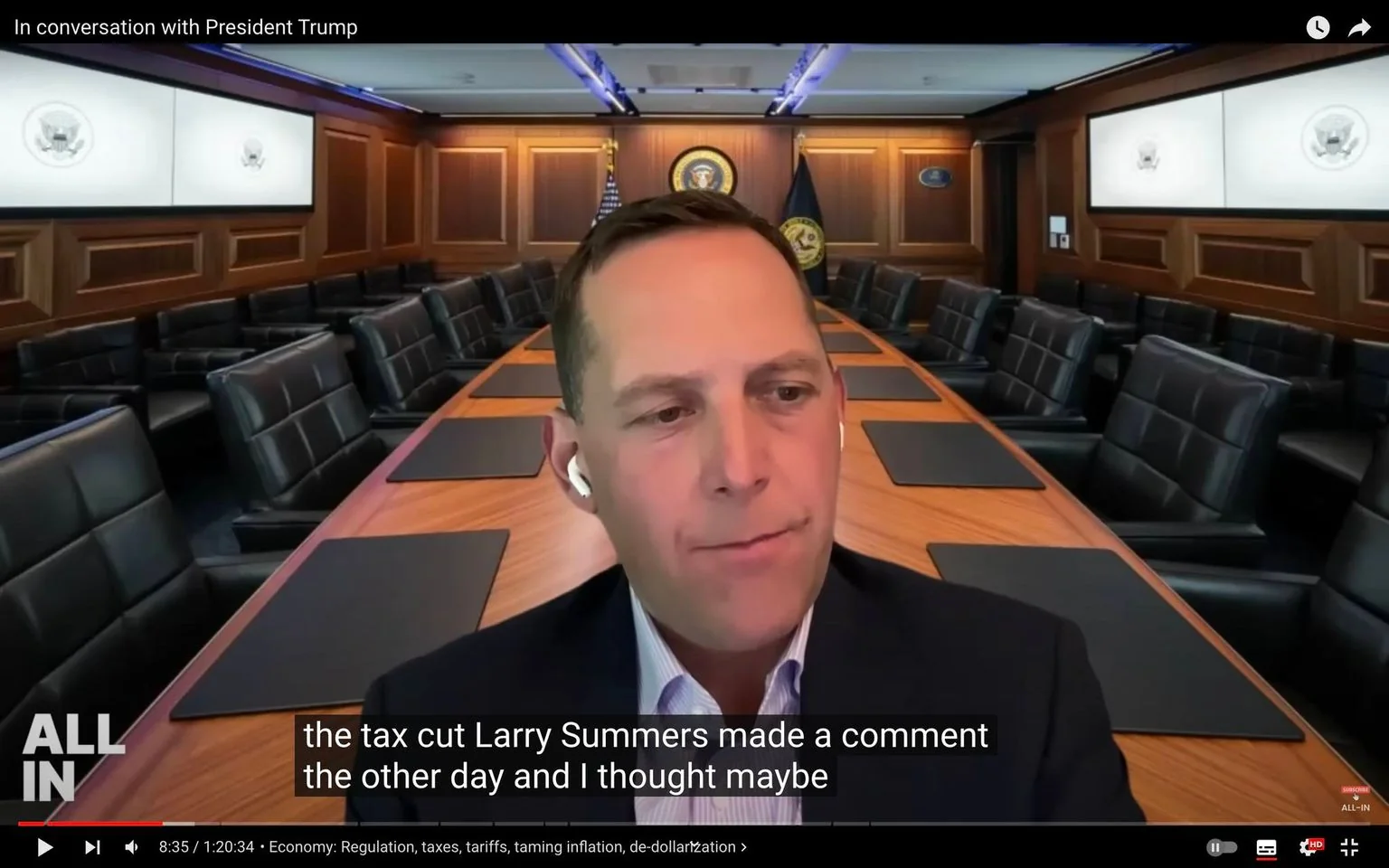 The image shows a man engaged in a virtual conversation, using a background that resembles a formal meeting room. The room is large and well-furnished, featuring a long conference table with black leather chairs. The setting includes wood-paneled walls and large monitors displaying official seals, suggesting a government or high-level business environment. The lighting is cool and professional, with a focus on creating an official atmosphere.
The man in the foreground appears to be mid-speech, wearing a dark suit and a light-colored shirt. He has earbuds in, indicating he is likely participating in an online call or webinar. His expression is attentive and engaged. The video title at the top reads "In conversation with President Trump," suggesting that the content involves a discussion or interview with the former president. Subtitles at the bottom show a partial sentence mentioning Larry Summers and a discussion about tax cuts. The video is titled "In conversation with President Trump" and is associated with topics such as the economy, regulation, and de-dollarization.