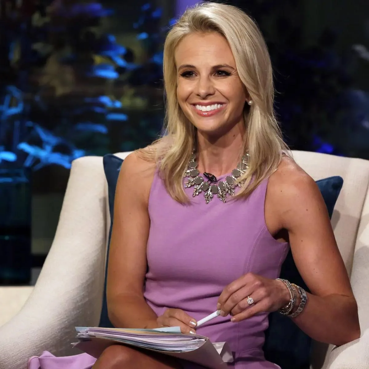 Elisabeth Hasselbeck is pictured seated in a studio setting, likely for an interview or talk show segment. She is wearing a sleeveless lavender dress that complements her blonde hair, styled straight and loose. A statement necklace with shiny embellishments adds a touch of elegance to her outfit. Elisabeth is smiling warmly, exuding a friendly and confident demeanor. Her posture is relaxed yet professional, as she holds a stack of papers and a pen, suggesting that she might be preparing to discuss or present something. The background is dimly lit with hints of blue and purple lighting, creating a sophisticated and intimate atmosphere typical of a television studio. The seating appears comfortable, with plush cushions, enhancing the inviting nature of the setting. There are no visible channel, title, or on-screen text elements, keeping the focus on Elisabeth Hasselbeck herself.