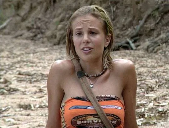 Elisabeth Hasselbeck is pictured in an outdoor setting that appears to be a beach or forest clearing, typical of a "Survivor" filming location. She is wearing an orange tube top with the "Survivor" logo prominently displayed, suggesting her participation in the reality TV show. A strap, likely from a bag, is slung over her shoulder. Elisabeth's expression is animated, possibly caught mid-conversation or during an interview segment. Her hair is pulled back, and she wears a necklace, adding to her casual, survivalist look. The background features a sandy, earthy terrain and some scattered branches, emphasizing the rugged, natural environment. The lighting suggests it is daytime, lending a realistic setting to the scene. The image captures the essence of a "Survivor" contestant in a candid moment, highlighting the reality competition's adventurous and challenging atmosphere.