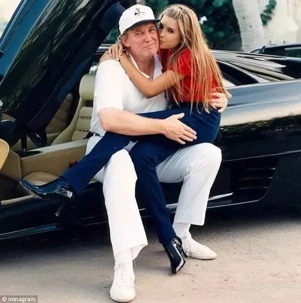 Ivanka and Trump are featured in this outdoor photograph. Trump, wearing a white polo shirt, white pants, and white sneakers, sits on the edge of a sleek black car with its door open. Ivanka, as a child, is dressed in a red top and blue pants. She is sitting on Trump's lap, wrapping her arms around him affectionately, and giving him a kiss on the cheek. The setting appears to be during the day, likely in a sunny, palm tree-lined location, adding to the casual and warm atmosphere of the image. The car's leather interior and the polished black exterior convey a sense of luxury. The photograph captures a candid and tender moment between the two, reflecting a familial bond. The lighting is bright and natural, highlighting the subjects and the car.