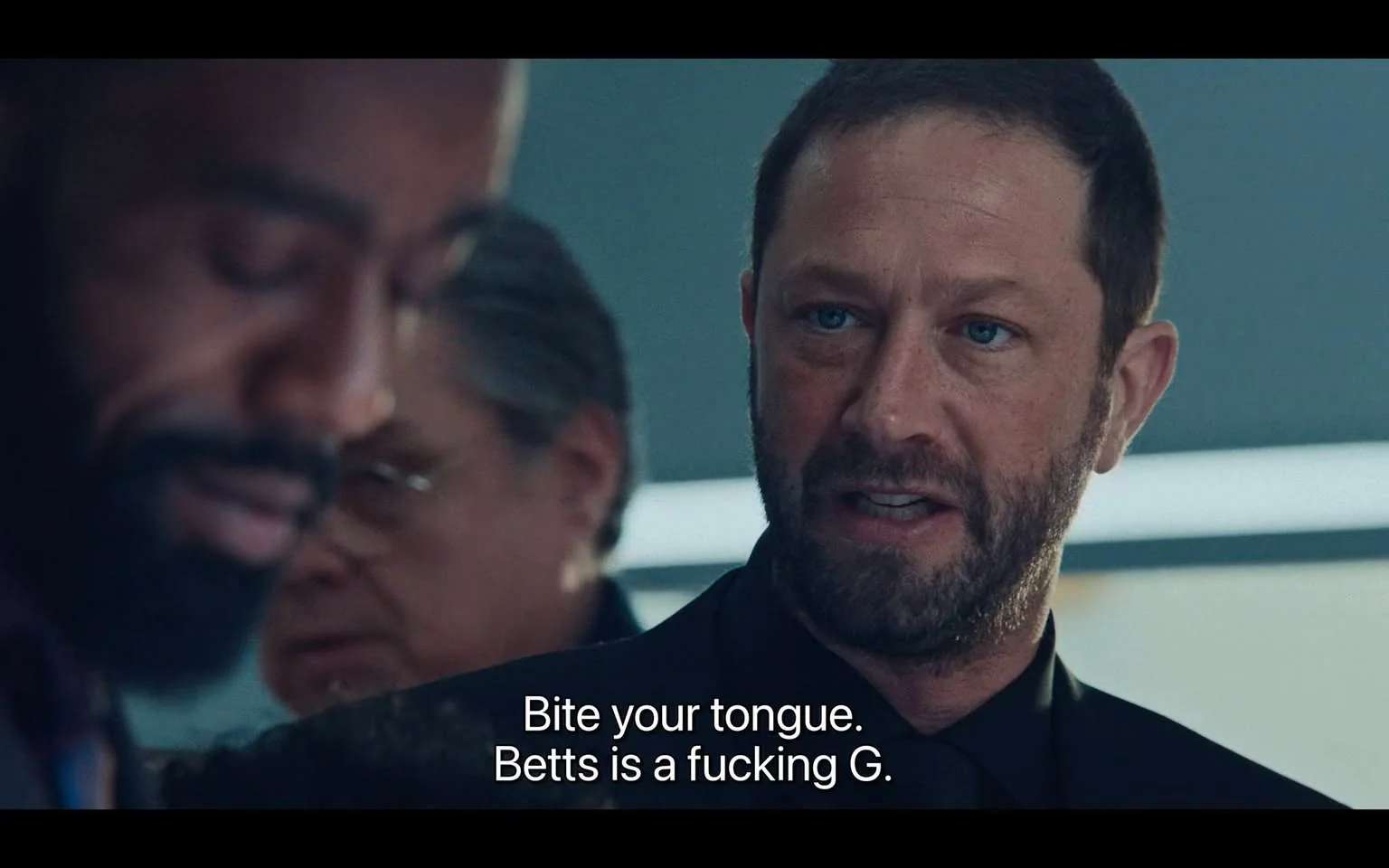 The image is a close-up scene from a film or TV show featuring a tense conversation between characters. The main focus is on a bearded man with short hair, who is speaking assertively. His expression is serious, and he is dressed in a dark shirt, suggesting a formal or professional setting. Beside him, there are two other figures partially visible, one with a beard and another in glasses. The lighting is neutral, with a soft focus on the main speaker, highlighting his facial features and intense gaze. The background is slightly blurred, emphasizing the characters in the foreground. The subtitle reads, "Bite your tongue. Betts is a fucking G.," indicating a conversation likely involving respect or status. The scene likely takes place indoors, possibly in an office or meeting room, given the formal attire and serious tone.