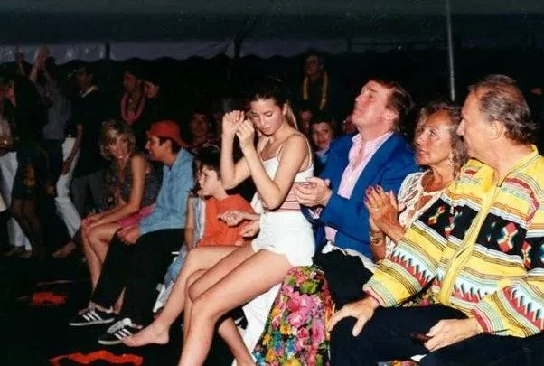 The image shows a group of people seated in what appears to be an indoor event setting, possibly a performance or show, given the audience-like arrangement. In the center of the image, Ivanka is sitting with others, wearing a white top and a short white skirt. She appears to be clapping or engaged in the performance, suggesting a lively or enthusiastic atmosphere. Next to her is Trump, dressed in a blue blazer and looking similarly engaged with the event.
The lighting is relatively dim, typical of event venues, with a tent-like structure visible in the background. The audience is arranged in rows of chairs, with a diverse group of people, including a man in a colorful patterned jacket to the right, contributing to the vibrant scene. Other attendees, including a child and a person in a red cap, are casually dressed, indicating a relaxed, informal gathering.
Overall, the photograph captures a moment of participation and enjoyment among the attendees, with Ivanka and Trump as central figures in this lively setting.