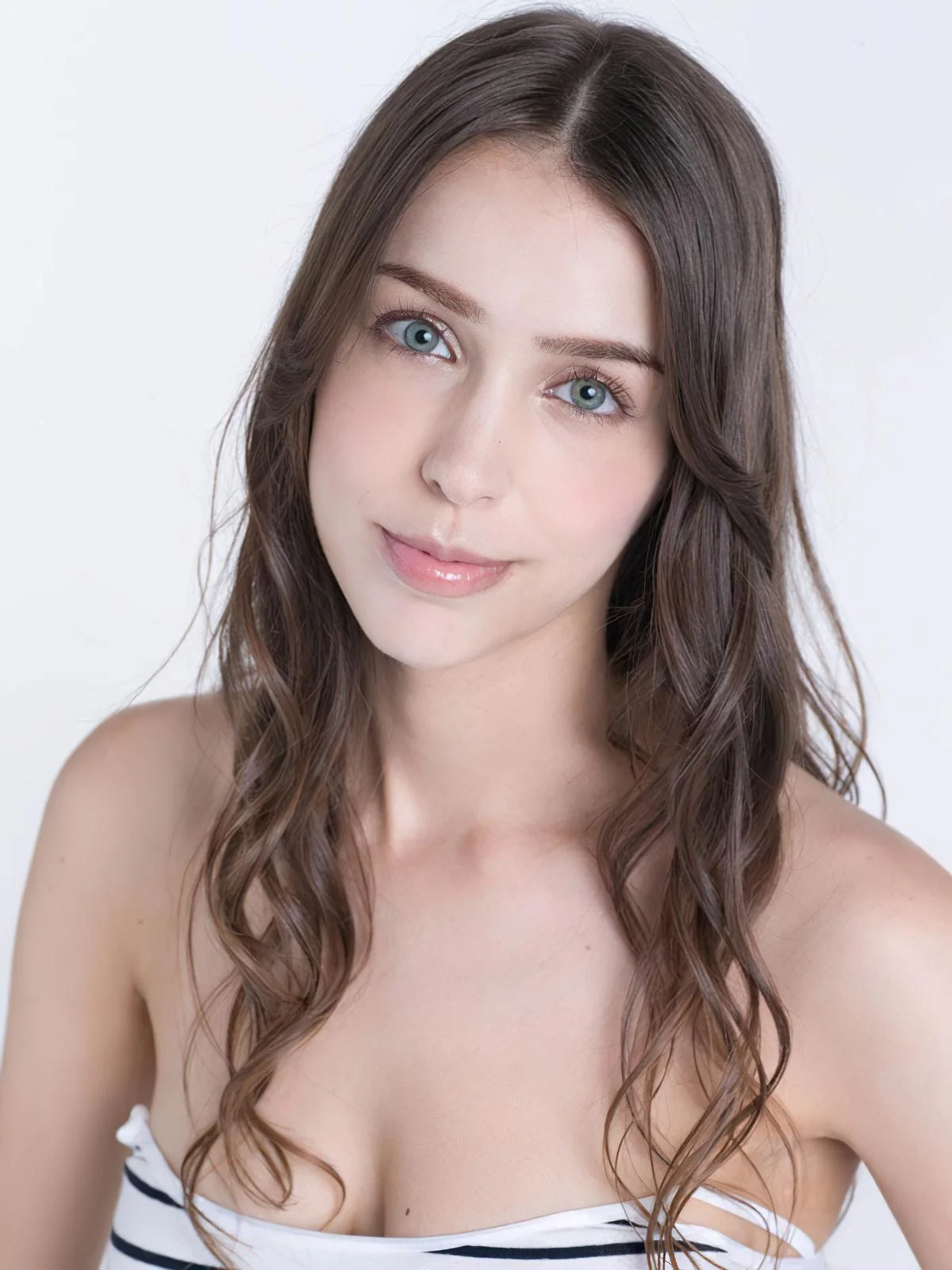 The image is a studio portrait featuring Stefanie Joosten. She is positioned against a plain white background, which accentuates her features prominently. Her long, wavy brown hair cascades softly over her shoulders. Stefanie is wearing a strapless top with black and white stripes, adding a subtle touch of style to the composition. Her expression is calm and poised, with a gentle smile that conveys a sense of warmth and friendliness. Her blue eyes are striking and complement the overall soft lighting of the scene, which highlights her complexion and creates a serene atmosphere. The image is likely a professional headshot, focusing on clarity and simplicity to emphasize her natural beauty. There is no visible text or branding in the image.