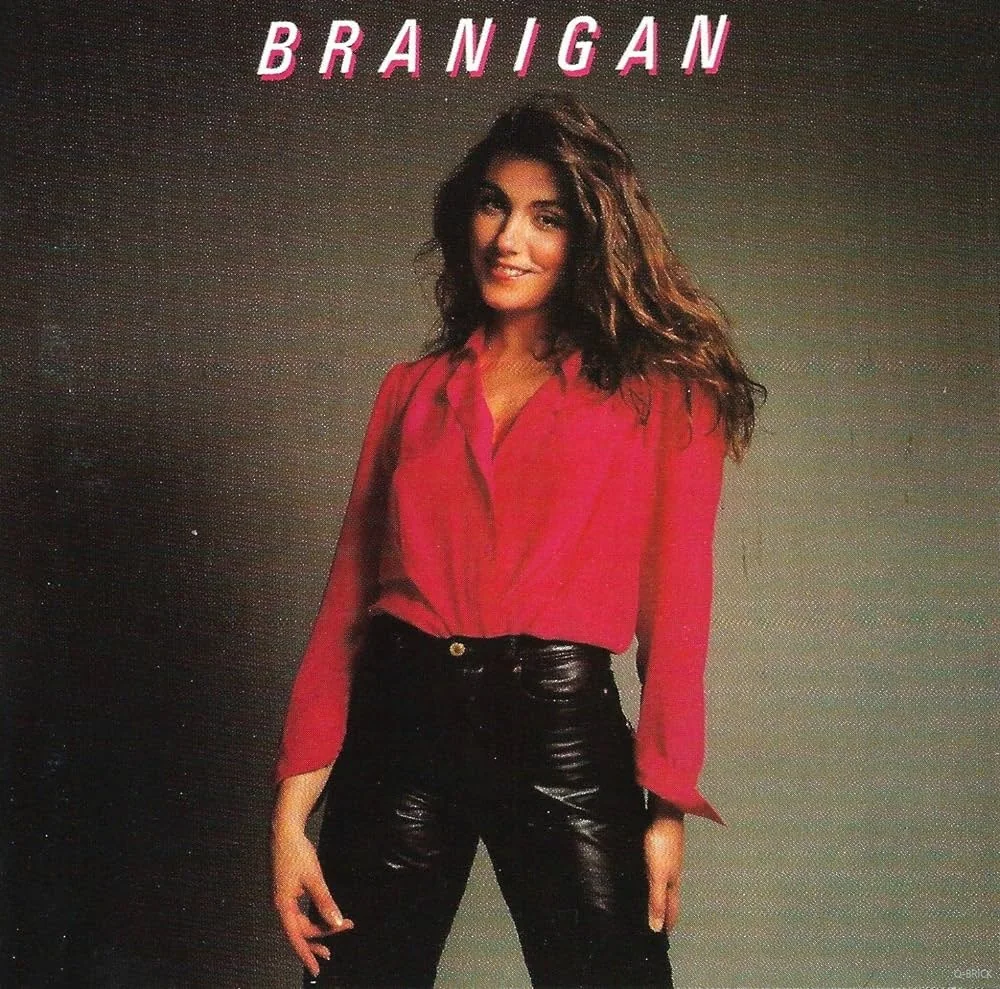 The image features a studio portrait of Laura Branigan, captured for an album cover. She is standing against a plain background, exuding a confident and approachable demeanor. Her outfit consists of a vibrant red blouse tucked into high-waisted black leather pants, embodying a classic 1980s style. Her hair is styled in voluminous waves, contributing to the overall dynamic and glamorous look. The lighting is even, highlighting her features and the textures of her attire.
The text "BRANIGAN" is prominently displayed at the top in bold, white capital letters with a slight shadow effect, enhancing the visual impact. The simplicity of the background and the focus on her attire and expression suggest an emphasis on the artist's persona and style. This image embodies a typical album cover aesthetic of the era, focusing on the artist as a central figure.
- Title: "BRANIGAN"