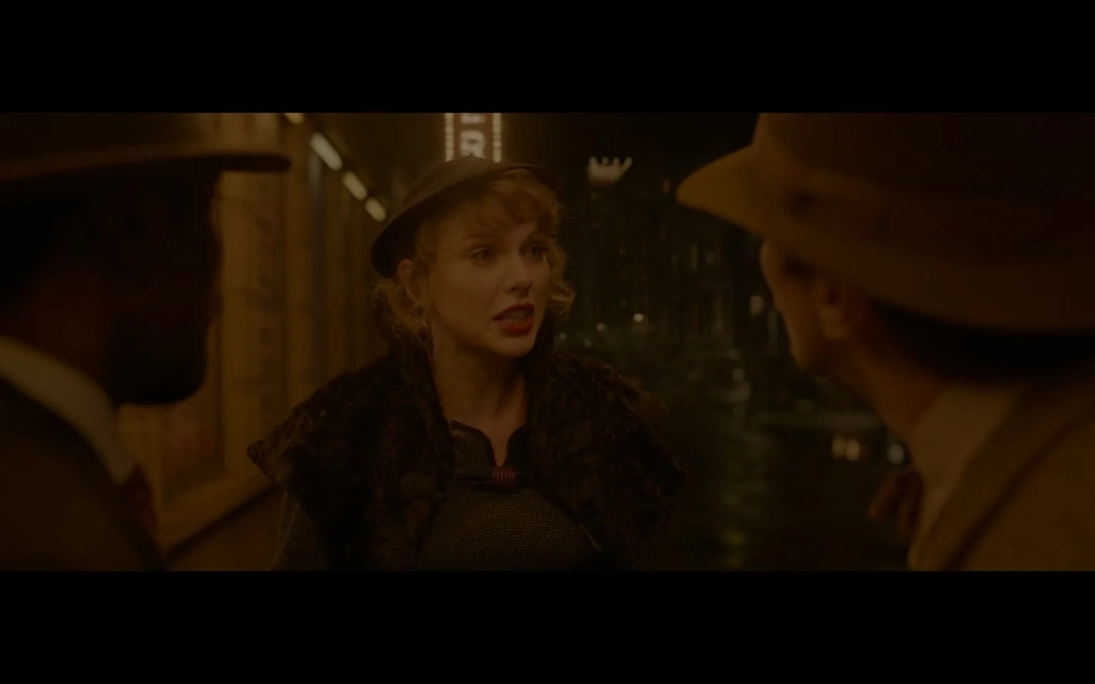 ```
Taylor Alison Swift is featured in this film scene set during the night. She stands centrally with two men on either side, engaging in what appears to be an intense conversation. The lighting is dim, contributing to a vintage atmosphere that complements the characters' period-appropriate attire. Taylor wears a dark, fur-trimmed coat and a hat, her expression serious and engaged, suggesting a dramatic moment. The scene is likely set in a city street, with blurred lights and buildings forming the background, enhancing the mood of the era being depicted. The two men are dressed in suits and hats typical of the time period, adding authenticity to the scene. The overall color palette is warm and muted, consistent with the nighttime setting.