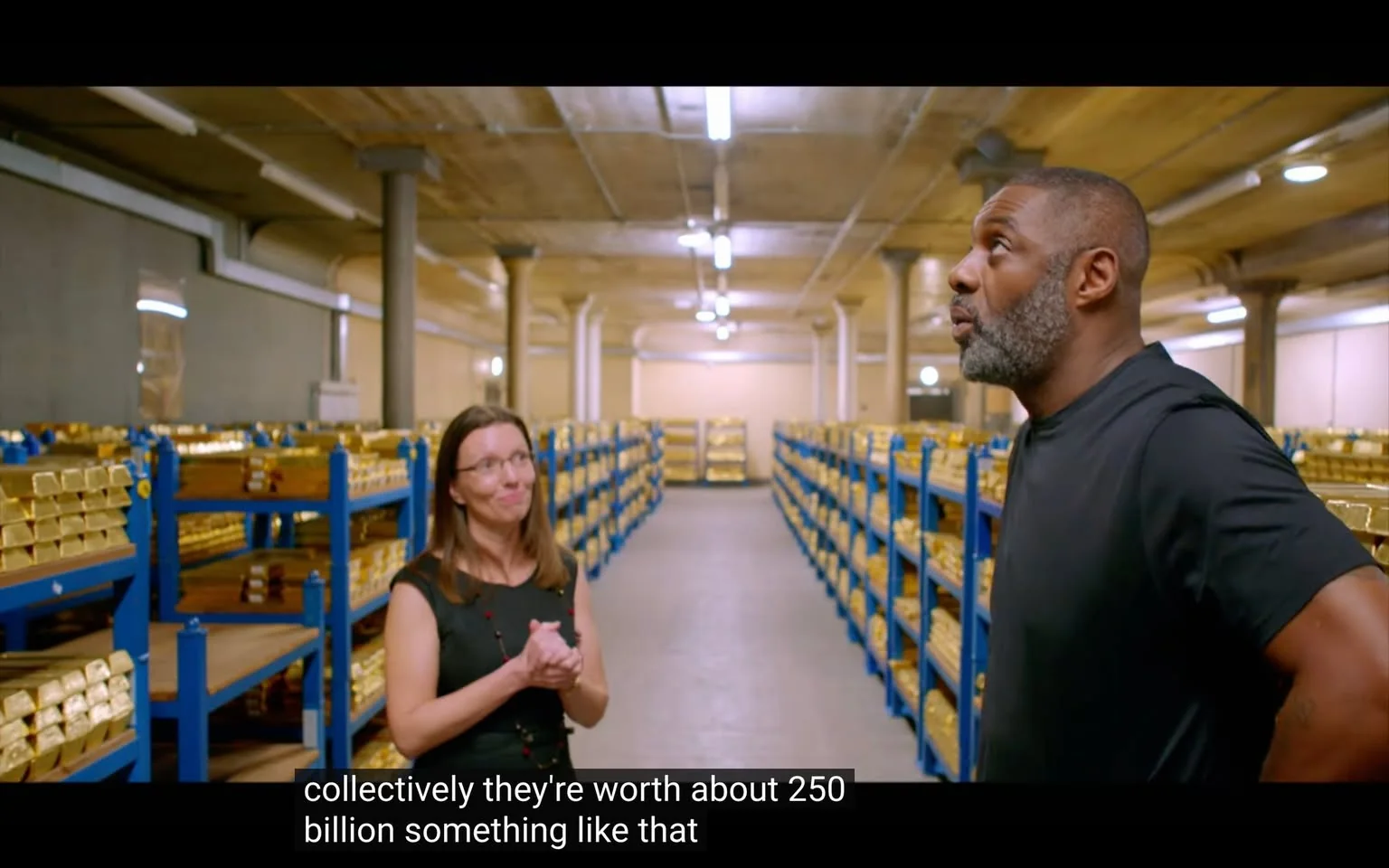 This image depicts an indoor scene in what appears to be a vault or secure storage area. The setting features numerous racks filled with gold bars, arranged neatly in rows, highlighting the immense wealth stored within. The area is characterized by sturdy columns and a ceiling with visible pipes, suggesting a basement or underground location.
In the foreground, two individuals are engaged in conversation. On the left, a woman with long brown hair, glasses, and a black dress appears to be explaining something, as indicated by her gesture and attentive expression. On the right, a man with a beard and short hair, wearing a black shirt, listens intently. The mood seems professional and focused, underscored by the serious yet calm expressions of both individuals. The lighting is bright and evenly distributed, illuminating the rows of gold, which adds to the scene's richness.
Subtitles at the bottom of the image reveal part of their conversation, mentioning the collective worth of the gold, estimated to be around 250 billion. This detail highlights the scale of wealth stored in the depicted location.