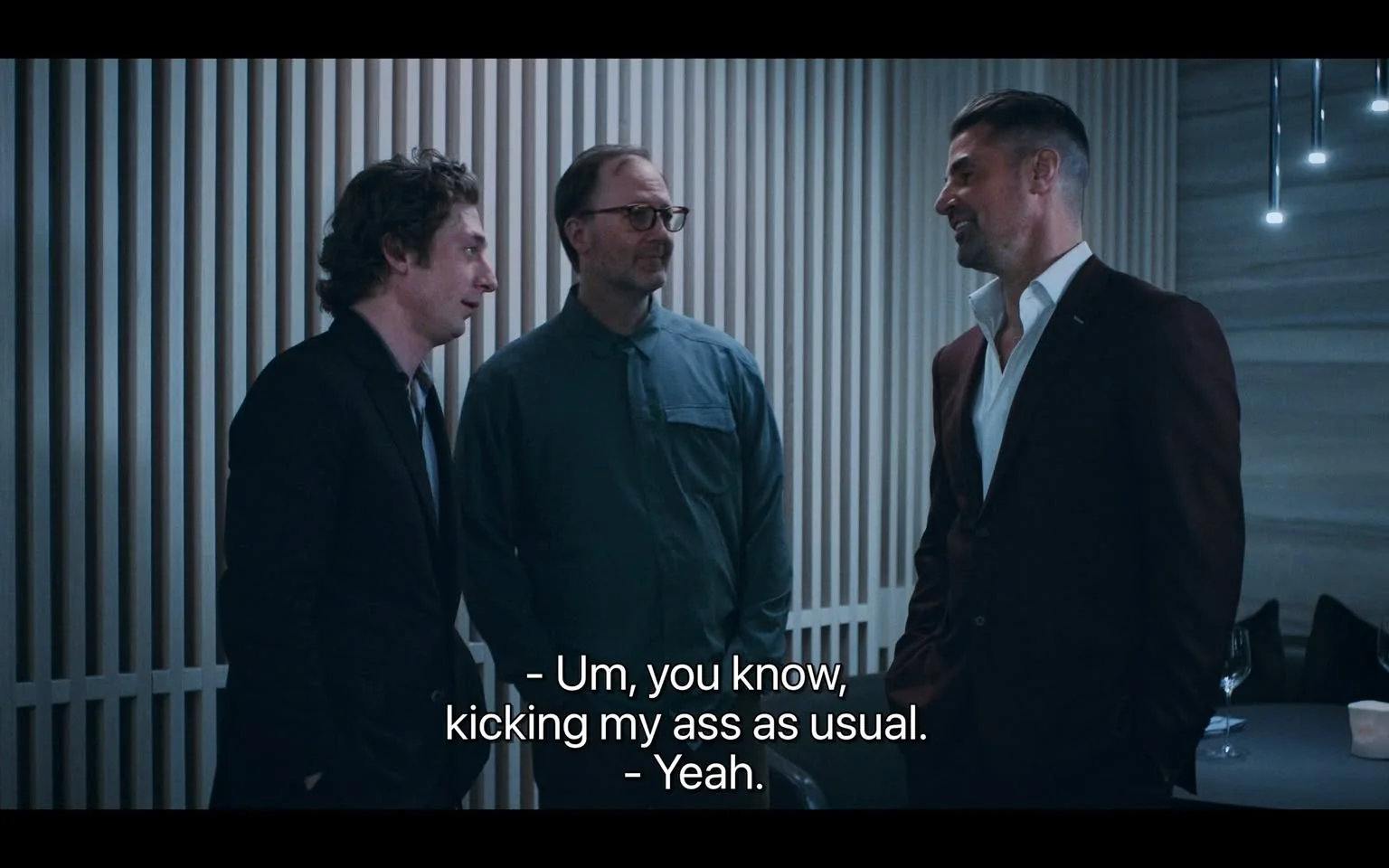 The image shows three men engaged in conversation in a modern, indoor setting, possibly in a dining or social area given the presence of wine glasses on a nearby table. The man on the left is wearing a dark suit with a tie, while the man in the middle has glasses and a button-up shirt. The man on the right is dressed in a dark suit with an open-collared white shirt, giving a casual yet sophisticated appearance. The background features a stylish, vertical striped wall, and the lighting is soft, suggesting an evening or intimate setting. The mood appears relaxed and informal, as indicated by the casual posture and subtle smiles of the men. The subtitles at the bottom of the image read, "- Um, you know, kicking my ass as usual. - Yeah." This exchange suggests a lighthearted or humorous conversation.