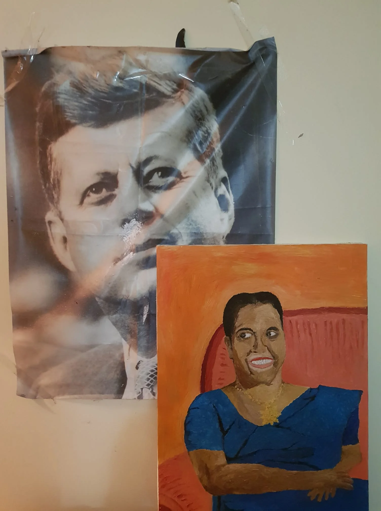 The image features two distinct pieces of art displayed on a wall. On the left, there is a black-and-white photograph of John F. Kennedy, showing him in a classic and thoughtful expression. The photograph has been affixed to the wall with tape, which is visible at the corners, giving it an informal presentation. To the right, there is a colorful painting depicting a woman in a blue dress seated against a bright orange background. The woman, identified as Milina Sumathipala, appears cheerful, with an expressive smile and relaxed posture. The contrast between the photograph and the painting is striking, emphasizing the different artistic mediums and styles. The lighting in the room is warm, casting a cozy ambiance that enhances the vivid colors of the painting. Both pieces are positioned closely, creating an intriguing juxtaposition between historical and personal significance. There are no visible identifying marks or text on the wall other than the artworks themselves.