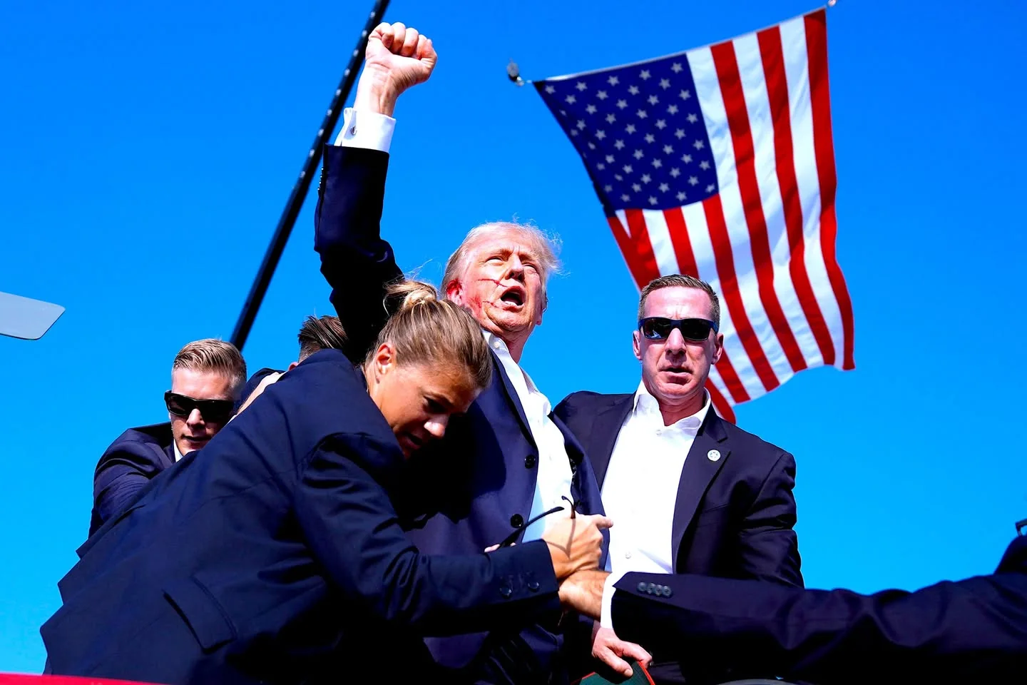 Donald Trump is featured prominently in this outdoor image. He is surrounded by several individuals, likely security personnel, wearing dark suits and sunglasses, indicative of a formal event or rally. The image captures a moment of high energy, with Donald Trump raising his fist in the air, suggesting a gesture of triumph or enthusiasm. The background is a clear blue sky, highlighting a large American flag which adds a patriotic element to the scene.
The security personnel appear vigilant, with one person notably leaning forward, possibly in the process of protecting or assisting Trump. The lighting is bright, casting clear shadows and emphasizing the vivid colors of the flag. The overall mood is dynamic, capturing a moment of action and intensity, typical of a political rally. No text or branding is visible in the image.