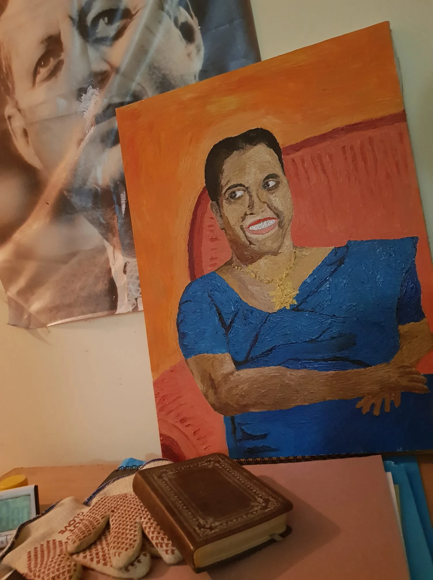 SECTION 2: Rich prose with relevant info
1) Rich description (8–12 sentences): The image shows a room containing a painting and a poster. The painting depicts a woman, identified as Milina Sumathipala, seated in a chair. She is wearing a blue dress and a gold necklace, with a broad smile on her face. The background of the painting is a warm orange color, creating a vibrant contrast with her dress. Next to the painting, there is a black-and-white poster of JFK, which captures a close-up of his face and adds a historical element to the scene. Below these artworks, various personal items are scattered, including a pair of patterned gloves, a small leather-bound book, and a stack of papers. The gloves have red accents, suggesting they might be for gardening or general work. The overall setting appears to be personal and intimate, likely belonging to someone with an appreciation for both history and family. The lighting is warm, highlighting the textures in the painting and the details of the objects.
2) On-screen text block:
- Title: "..."
