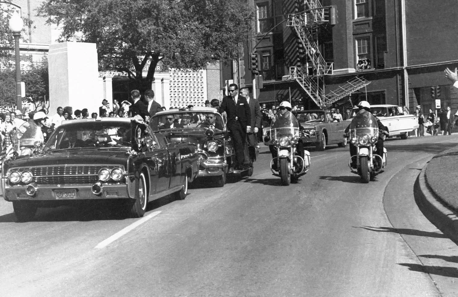 This black and white image captures a historic motorcade proceeding through a city street. In the foreground, a large, convertible automobile is prominently featured, flanked by two police motorcycles. The car is part of a procession, moving in formation along a gently curving road. The mood is formal and ceremonial, with the policemen wearing helmets and the drivers dressed in suits, reminiscent of a significant political or state event from the 1960s.
Spectators can be seen lining the sidewalks, watching the vehicles pass by, while a few people are visible in an adjacent building, observing from windows. The backdrop includes trees and a brick building with visible fire escape ladders. Text on the license plate of the lead car, marked with "GG 300," suggests an official designation. The lighting is bright and clear, indicating a sunny day, which adds to the vividness of the scene. The composition emphasizes the sense of movement and importance of the event.