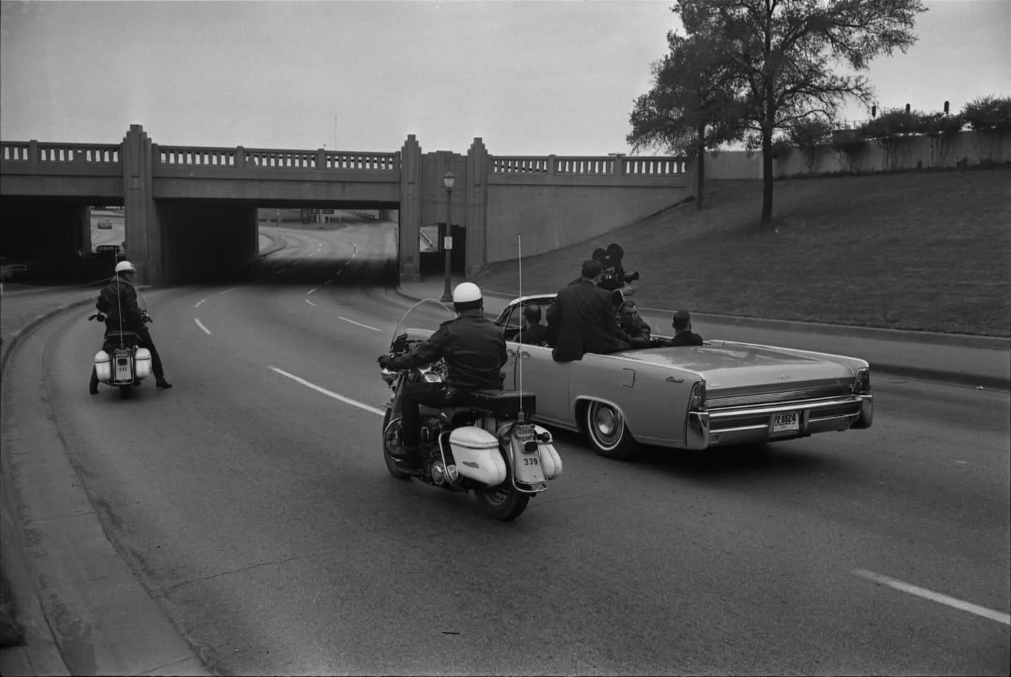 The image shows a monochrome photograph depicting a motorcade taking place on a road next to a grassy area with a few trees. In the foreground, there are two police officers on motorcycles escorting a convertible car. The car is notable for being a Lincoln Continental, which is a classic and recognizable model. Inside the vehicle, several people are visible, including a driver and passengers, with one person standing and handling a large camera, suggesting they are documenting the event. The convertible has the top down, allowing a clear view of its occupants.
The road is lined with street lamps, and in the background, a concrete overpass stretches across the road. The weather appears overcast, with soft lighting and no harsh shadows. The scene evokes a sense of historical significance, possibly related to a well-known event or figure, given the formal attire of the individuals and the presence of the police escort. The composition is balanced, with the road curving gently to the left, leading the viewer's eye through the scene. The license plate on the vehicle reads "PZ 8524," and the motorcycles have different identification numbers visible on their rear fenders.
