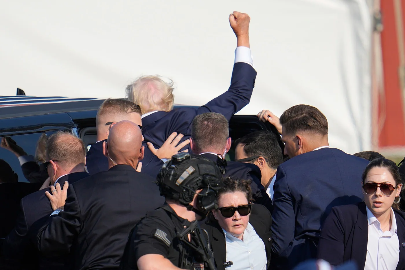 Donald Trump is seen in the image surrounded by security personnel as he is getting into a vehicle. The setting appears to be outdoors, likely at an event or public appearance. Donald Trump is partially visible, mostly from the back, as he raises his right arm with a clenched fist. He is wearing a dark suit and his distinctive hair is visible. The security team around him is dressed in dark suits, with some wearing sunglasses and earpieces, indicating they are likely part of a protective detail. One of the security members in the foreground is wearing a tactical vest and looking towards the camera. The scene is bustling, with a sense of urgency and protection as the group moves towards the vehicle. The lighting is bright, suggesting it is daytime. The vehicle is a dark SUV, commonly used for transporting high-profile individuals. The overall mood conveys a mix of determination and protection in a public setting.