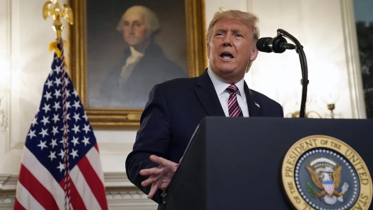 In the image, Donald Trump is speaking at a podium labeled "President of the United States." He is wearing a dark suit with a white shirt and a red striped tie. His expression appears focused and engaged as he gestures with one hand. Behind him, an American flag stands prominently, adding to the formal and patriotic setting. A portrait of George Washington, attributed to Gilbert Charles Stuart, hangs on the wall in the background, providing historical context to the scene. The lighting is bright, emphasizing the seriousness of the occasion. The setting suggests a speech or announcement in a formal governmental environment, likely within a significant building like the White House. The overall mood conveys authority and importance, underscored by the symbols of the American presidency and heritage.