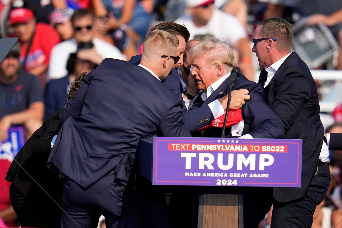 The image captures Donald Trump at a campaign rally event. He is surrounded by several men, who appear to be security personnel, as they hold and support him. Donald Trump is at a podium with a microphone, wearing a dark suit and a white shirt, partially obscured by the men around him. The podium has prominent signage displaying phrases like "TEXT PENNSYLVANIA TO 88022" and "TRUMP MAKE AMERICA GREAT AGAIN! 2024" in bold, uppercase letters. In the background, a crowd of people is visible, many wearing red, white, and blue clothing, indicative of a political rally setting. The scene is brightly lit, suggesting a daytime event, with a busy and dynamic atmosphere. The men around Donald Trump are dressed in suits, looking focused and intent on their task. The overall mood seems tense, possibly indicating an unexpected situation.
- Title: "TEXT PENNSYLVANIA TO 88022"
- Site / app: "..."