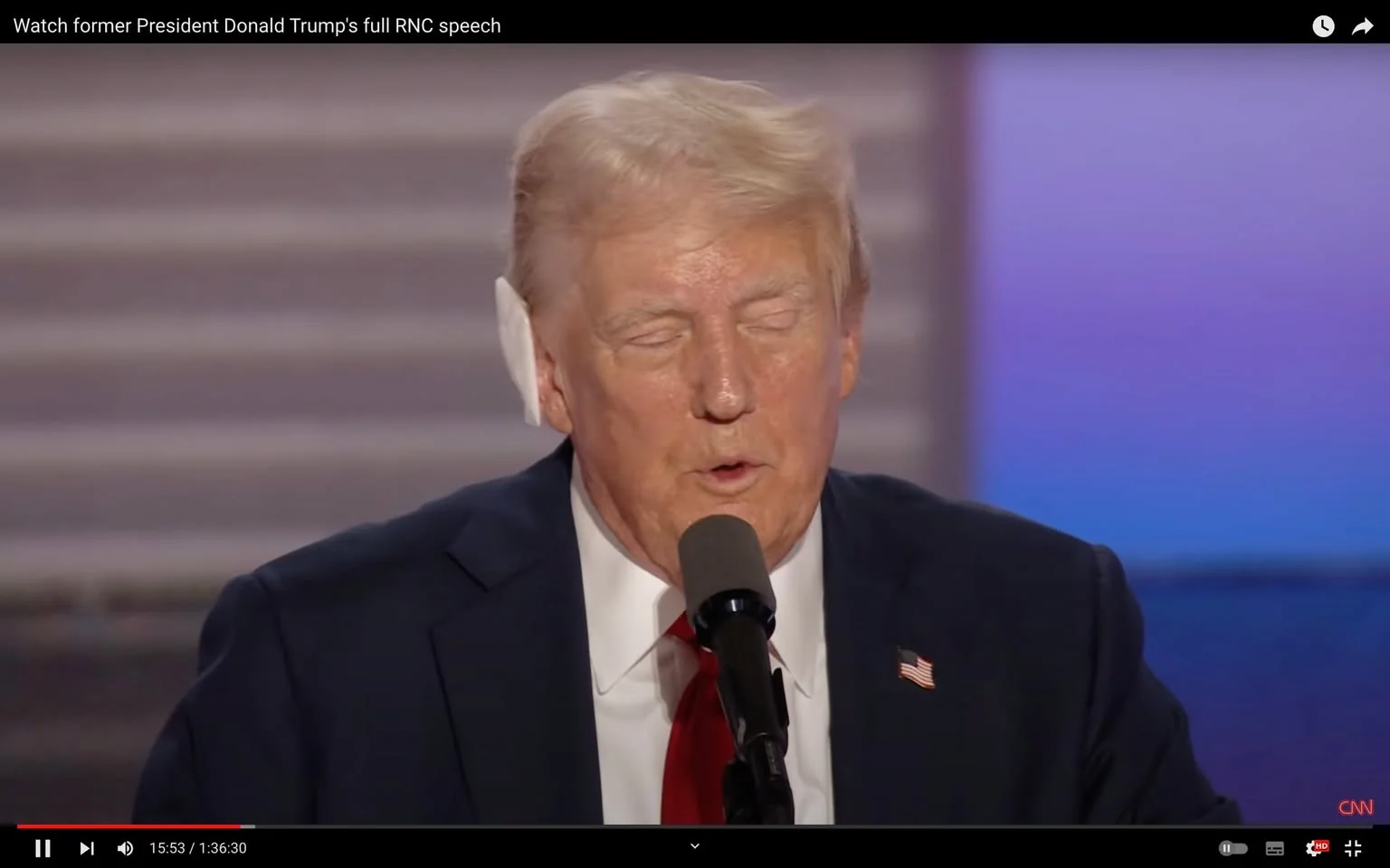 Donald Trump is depicted in this video frame during his full RNC speech, as indicated by the title text. He is standing at a podium with a microphone, wearing a dark suit, white shirt, and a red tie adorned with a small American flag pin on his lapel. The background features a soft gradient of purple and blue tones, creating a formal atmosphere typical of such events. Donald Trump appears to be speaking or about to speak, with his eyes closed and mouth slightly open, suggesting a pause or emphasis in his speech. The lighting is bright and focused on him, highlighting his facial features and attire. This frame is part of a broadcast on CNN, as shown by the logo at the bottom right of the image and within the title text itself. The setting appears to be indoors, consistent with a formal event or convention setting.