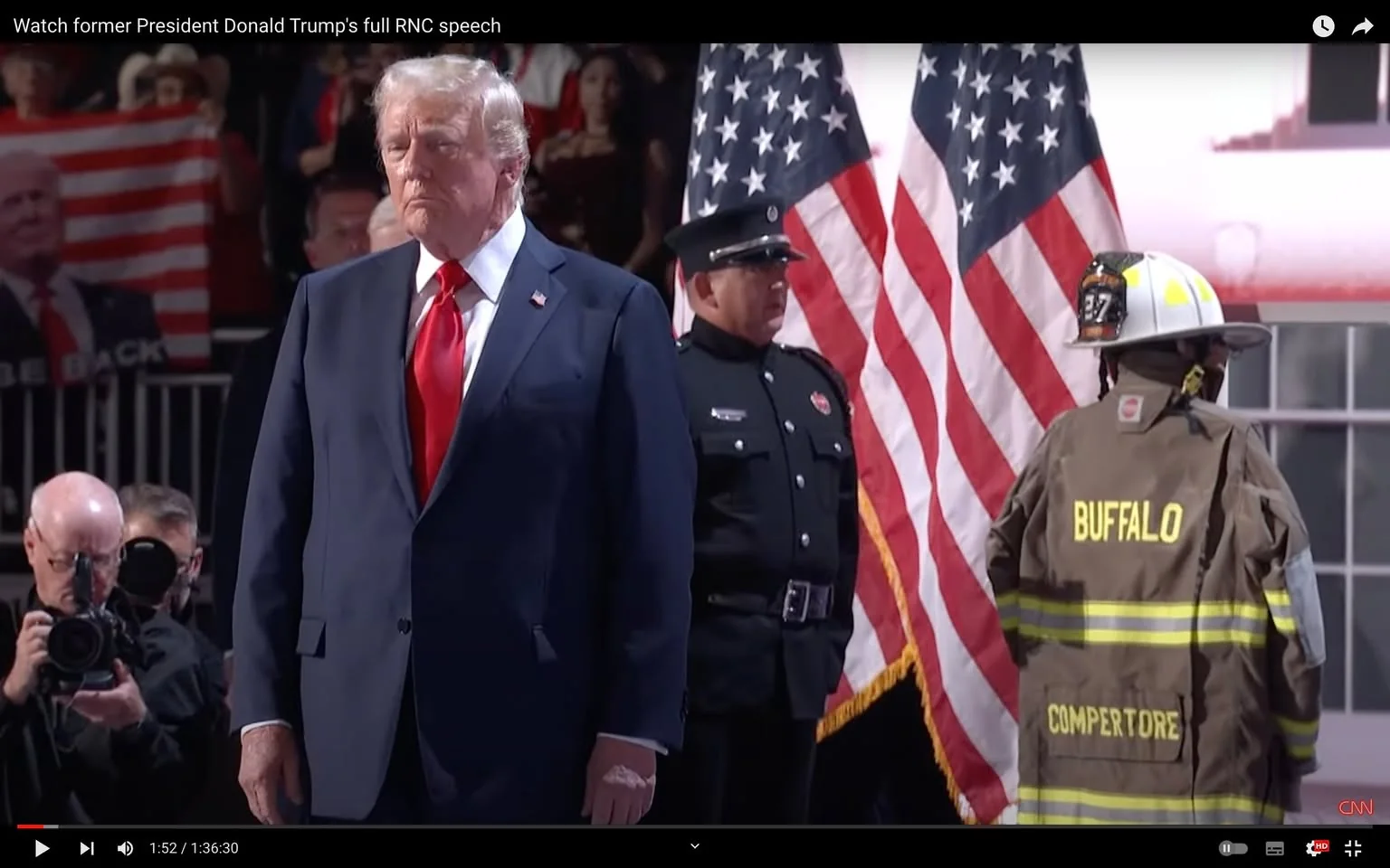Donald Trump is seen standing on a stage, wearing a navy blue suit with a white shirt and a bright red tie. His expression is serious and he appears to be attending a formal event. Behind him, two American flags are prominently displayed, symbolizing the patriotic setting of the event. To his right, a person in a firefighter's uniform with "BUFFALO" and "COMPERTORE" written on the back is visible, indicating a tribute or honor to emergency services. Another person dressed in a formal black uniform stands to the left, enhancing the formal atmosphere.
The background is filled with an audience, some of whom are taking photographs, and there is a visible poster with an image of Donald Trump and an American flag pattern. The lighting is bright, focusing on the stage area where the main event is taking place. The image is a video frame from a recording, as suggested by the play bar at the bottom and the YouTube interface elements. The overall mood is solemn, with an emphasis on national pride and civic duty.
- Title: "Watch former President Donald Trump's full RNC speech"
- Channel / profile: "CNN"
- Site / app: "YouTube"