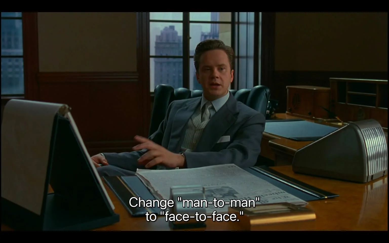 ```
The image is a scene from a film set in an office. A man in a formal suit sits at a desk, suggesting a business setting. He appears to be engaged in a conversation, as indicated by his hand gesture and the subtitle accompanying the image: "Change 'man-to-man' to 'face-to-face.'" This suggests an emphasis on modifying language for inclusivity or accuracy. The office is elegantly furnished, with a large desk, leather chairs, and a window view showing tall buildings, indicating a cityscape outside. The light is natural, implying daytime. The presence of a newspaper on the desk suggests the character might be in a position of authority or involved in current events. The atmosphere is professional, with muted colors enhancing the formal tone.