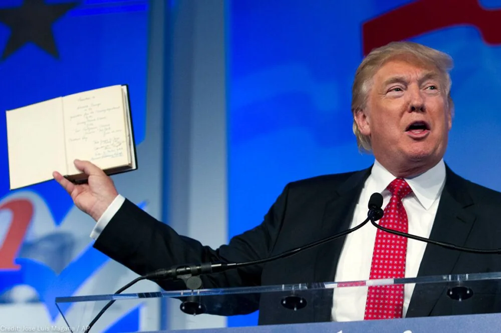 Donald Trump is depicted in the image, standing at a podium, appearing to deliver a speech or presentation. He is holding an open book in his left hand, displaying written notes or a list to the audience. The setting is likely an indoor event, as indicated by the presence of a microphone and a backdrop featuring graphical elements. Donald Trump is dressed formally in a dark suit, white shirt, and a red tie, which is typical of his public appearances.
The background features a blue hue with abstract designs, possibly reflective of an event or conference theme. Donald Trump’s expression is animated, suggesting engagement with the audience or emphasis on a point he is making. The lighting is bright, focusing attention on him as the central figure in the scene. The book he holds contains handwritten text, which includes menu items such as "Grilled Cheese & Tomato Soup" and "Grilled Veggie Wrap."
Overall, the image captures a moment where Donald Trump is actively communicating, possibly emphasizing or illustrating a point using the notes in the book.