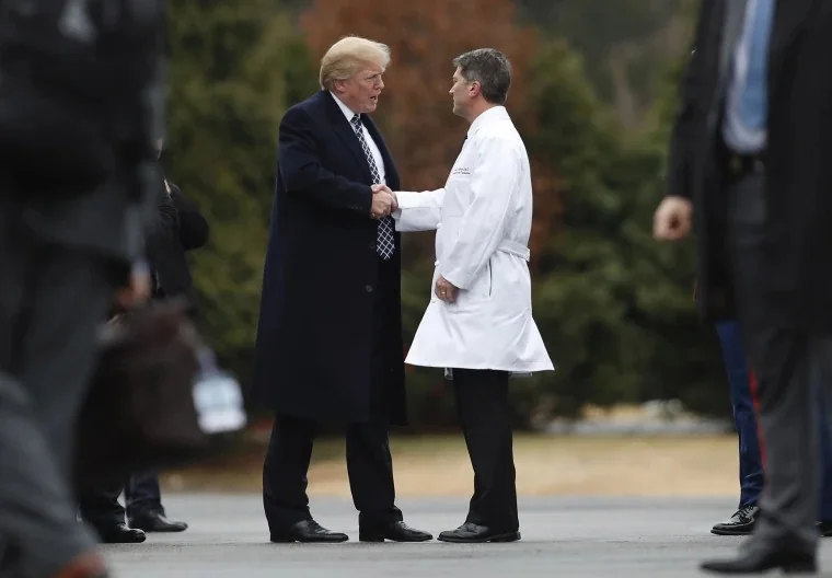 Donald Trump is seen in an outdoor setting, engaging in a handshake with a man wearing a white lab coat. The encounter takes place on what appears to be a tarmac or open area surrounded by trees. Donald Trump is dressed in a dark overcoat, a white shirt, and a patterned tie, exuding a formal and authoritative presence. His hair is styled in his signature manner, and he appears to be focused and attentive during the handshake.
The man in the lab coat, possibly a doctor or medical professional, stands respectfully as they converse. The background features blurred figures, likely security or staff, indicating a formal and possibly official event. The overall mood is professional, with both individuals maintaining composed expressions. The lighting is natural, suggesting daytime, and the setting is neutral, with muted colors that emphasize the formality of the interaction.