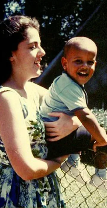 The image depicts a woman holding Toddler Barack Obama in her arms. The setting is outdoors on a sunny day, evident from the bright lighting and clear sky in the background. The woman, wearing a floral dress, is smiling and looking at Toddler Barack Obama, who is wearing a light blue shirt and pants. He appears joyful, smiling broadly at the camera. They are positioned near a chain-link fence, which is partially visible in the foreground. The greenery and blurred background suggest a park-like environment. The overall mood of the image is warm and affectionate, capturing a candid moment of happiness and connection between the two individuals. The photograph has a nostalgic quality, typical of family snapshots from past decades.
