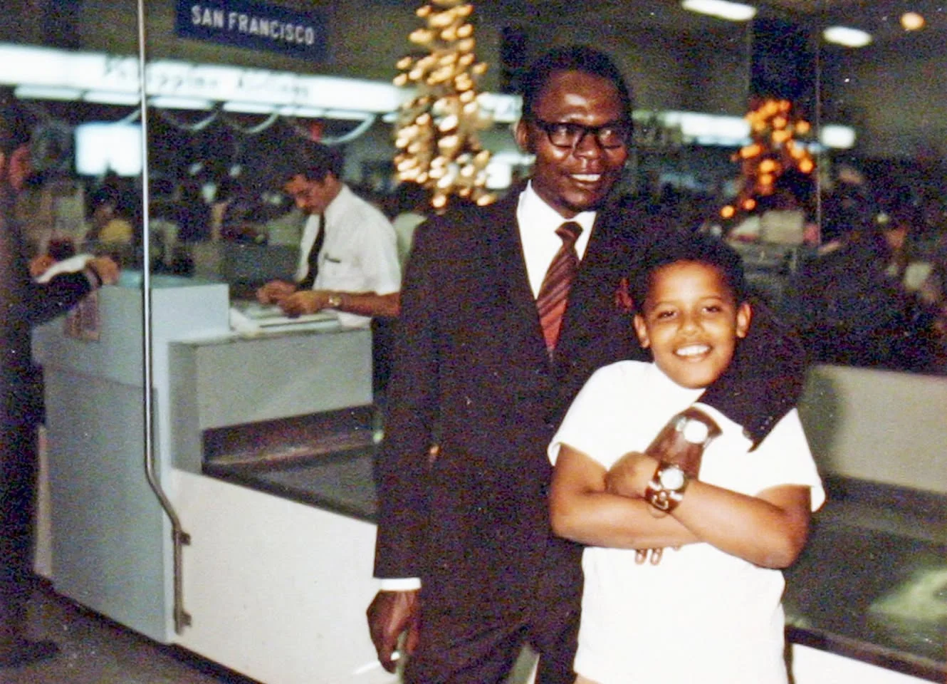 In this vintage photograph, Younger Barack Obama is seen embracing Barack Hussein Obama Senior. They are positioned in an airport setting, with signage indicating a connection to San Francisco visible in the background. The younger Obama, wearing a casual white t-shirt, is smiling broadly and holding onto a small object, possibly a toy or stuffed animal. Meanwhile, Barack Hussein Obama Senior is dressed in a formal dark suit, complemented by a tie and glasses, exuding a sense of warmth and pride as he has his arm wrapped around his son.
The background of the image features Christmas decorations, suggesting the photo might have been taken during the holiday season. Behind them, an airport counter and a staff member attending to paperwork can be seen, capturing a slice of daily life in this bustling environment. The lighting is typical of indoor settings from the era, giving the scene a nostalgic feel. The composition of the image focuses on the affectionate bond between father and son, highlighted by their contrasting attire and united expressions.