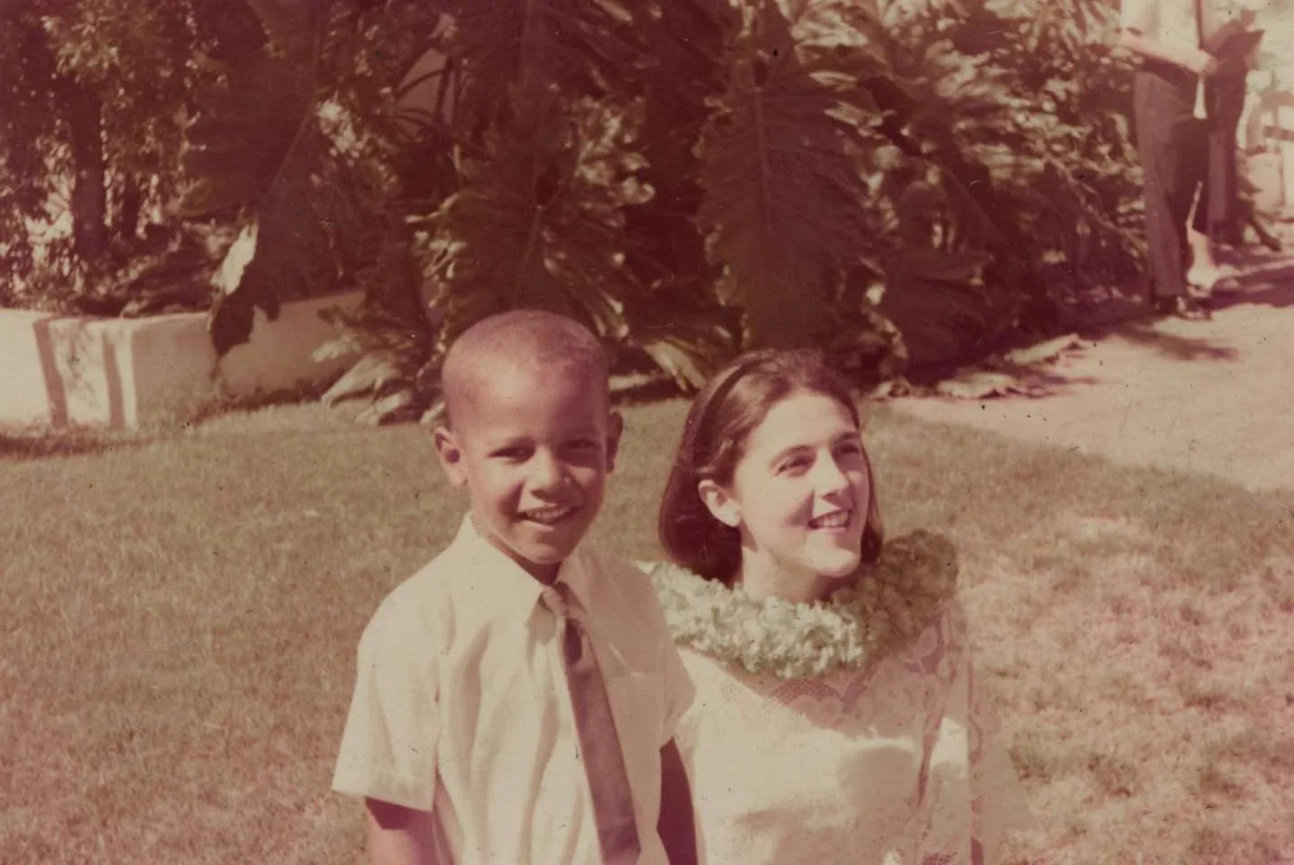 In this vintage photograph, Child Barack Obama is seen standing on a lawn next to a woman. The setting appears to be outdoors on a sunny day, characterized by lush greenery in the background, including large leafy plants. Child Barack Obama is wearing a light-colored shirt and a tie, exuding a sense of formality that contrasts with the casual outdoor setting. The woman beside him, who seems to be dressed for a special occasion, is wearing a delicate lace dress and a lei around her neck, suggesting a celebration or cultural event. Their expressions are warm and relaxed, hinting at a joyful interaction. The image has a slightly faded, sepia tone typical of older photographs, evoking a sense of nostalgia. In the background, another person can be vaguely seen, adding depth to the composition. Overall, this image captures a candid moment from Barack Obama’s childhood, reflecting both personal and cultural elements.