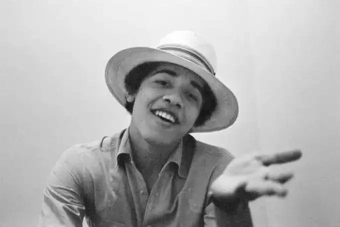 This black and white photograph captures a young Barack Obama in a relaxed and informal pose. He is seated indoors against a plain background, which emphasizes his presence in the frame. Barack Obama is wearing a light-colored button-up shirt, and he has a straw hat with a dark band adorning his head, adding a touch of casual style to the portrait. His expression is engaging and friendly, with a wide, open smile that exudes a sense of youthful exuberance and approachability. His left hand is slightly outstretched towards the camera, enhancing the sense of spontaneity and interaction with the viewer. The lighting is soft and even, typical of indoor settings, which helps in highlighting the subject's features without casting harsh shadows. The photograph, likely taken during his early years, offers a glimpse into the personal style and charisma that would later become synonymous with his public persona.