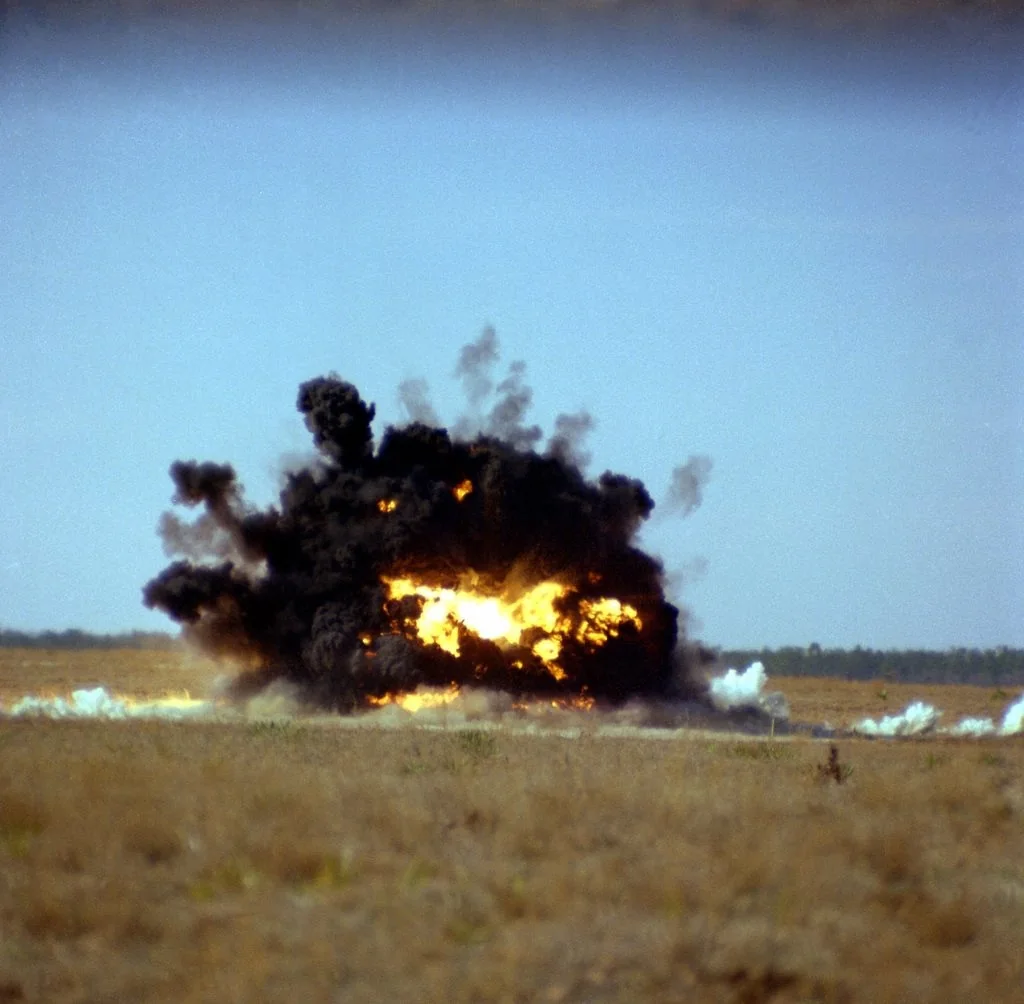 The image captures a dramatic explosion occurring in an open, arid landscape, likely a testing or training ground. In the center of the frame, a large fireball is engulfed in thick black smoke, indicating a powerful detonation. The bright yellow and orange of the flames contrast strikingly with the dark smoke, creating a vivid and intense scene. The ground appears dry and barren, with sparse vegetation visible in the foreground and a line of trees or distant brush on the horizon under a clear blue sky. The overall atmosphere suggests a controlled explosion, possibly for military or cinematic purposes, given the lack of human presence or structures in the immediate vicinity. The lighting is natural, with the sun illuminating the scene from above, emphasizing the billowing smoke and fiery core of the blast.