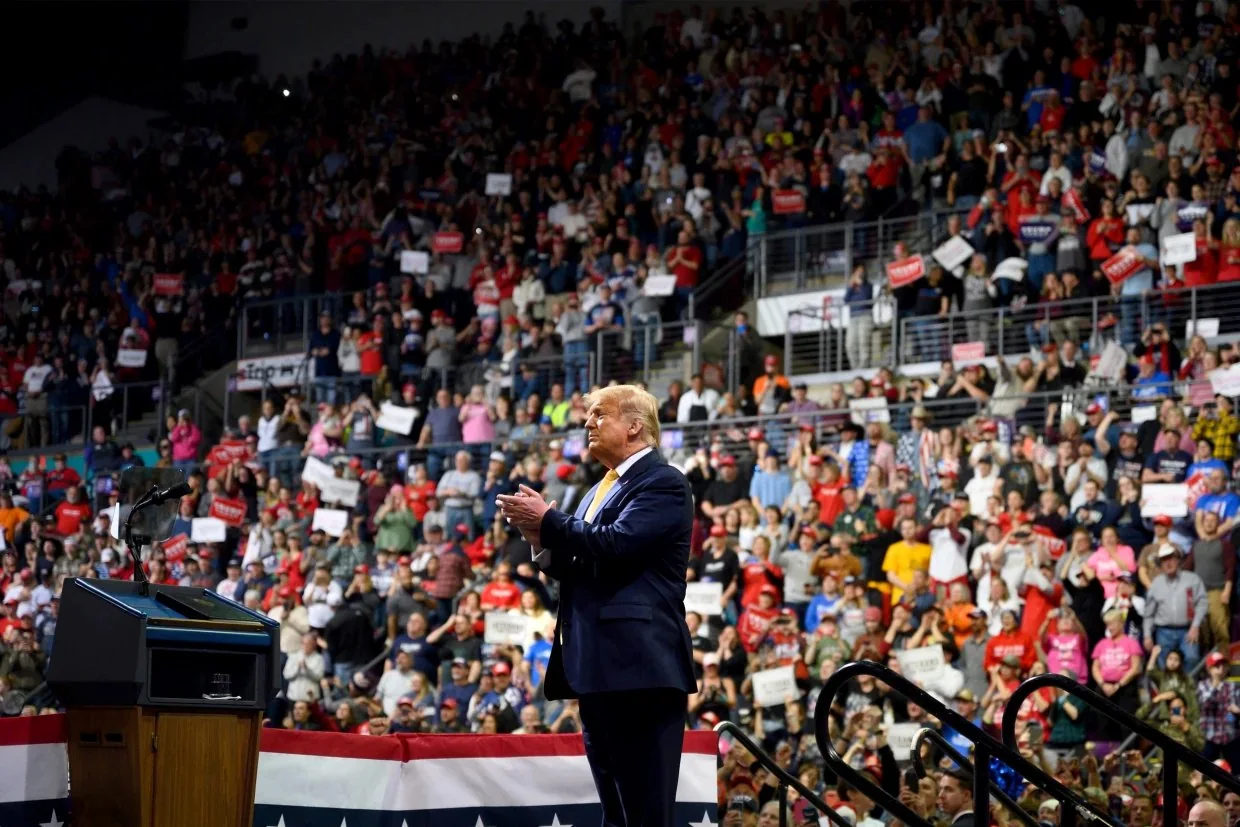 Donald Trump is seen at a large indoor rally, standing on a stage in front of a substantial crowd. The setting is a packed auditorium with multiple tiers of seating filled with people holding signs, many of which are red and white. The audience appears enthusiastic, with individuals wearing hats and shirts in various colors, including red, which is commonly associated with political rallies. Donald Trump is positioned in the foreground, facing the crowd, clapping his hands. He is dressed in a dark suit with a white shirt and yellow tie, looking engaged in the event. The stage is modestly decorated with a podium draped in a fabric featuring stars and stripes, echoing a patriotic theme. The lighting is bright, focusing on Trump and illuminating the cheering audience. The mood is energetic and lively, characteristic of a political rally with a significant turnout.