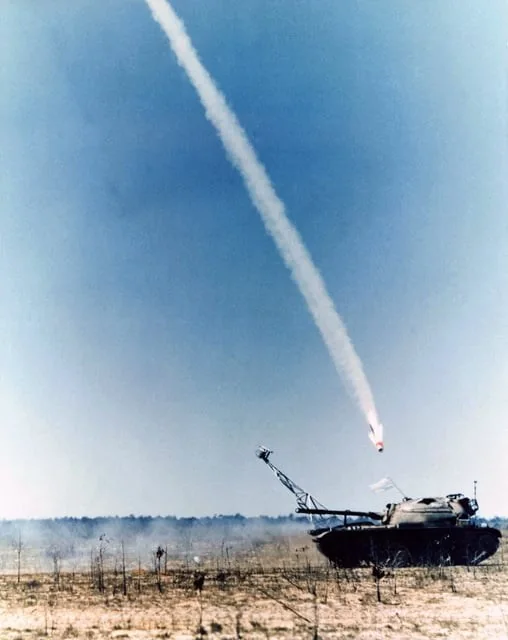 The image captures a dramatic scene of a missile launch from a military vehicle in a desert setting. The vehicle, which appears to be a tracked military platform, is positioned in the lower right corner of the frame. It is equipped with a missile launching system, which is actively firing a missile into the clear blue sky. The missile is at the peak of its trajectory, leaving a prominent white smoke trail that curves across the sky, indicating its high speed and power.
The terrain is dry and sparse, characteristic of desert environments, with faint vegetation visible. The lighting suggests it is daytime, with a clear sky providing a stark contrast to the smoke trail. The photograph's composition emphasizes the missile's upward motion, capturing the moment of propulsion and the technology involved in such military operations. The overall mood is one of tension and technological prowess, highlighting the vehicle's role in missile defense or attack scenarios.