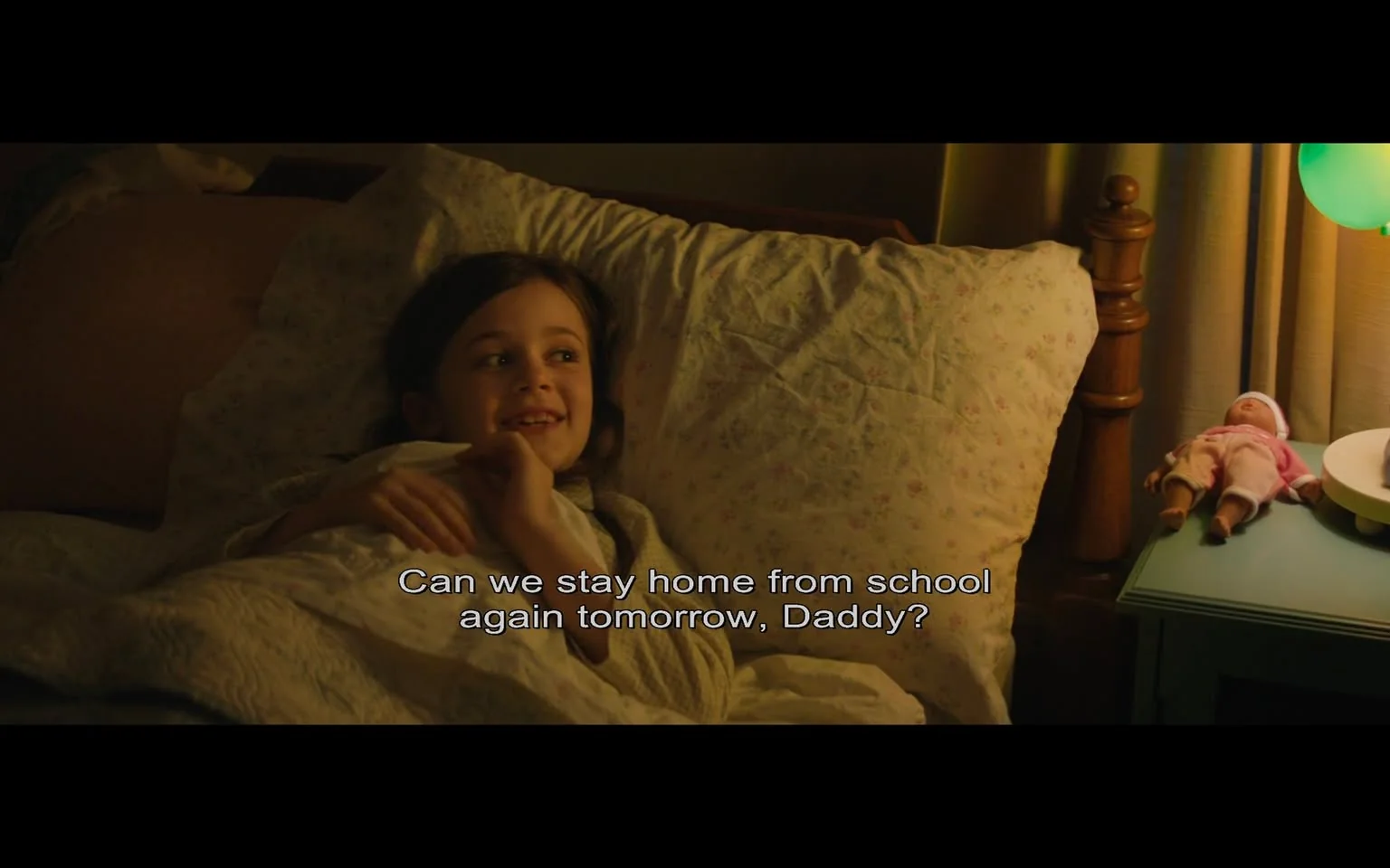 The image shows a young girl lying in bed, covered with a floral-patterned quilt. She appears to be in a cozy bedroom setting, likely at night, given the soft, warm lighting. The girl is smiling and looking upward, suggesting she is speaking to someone nearby, possibly her father, as indicated by the subtitle, "Can we stay home from school again tomorrow, Daddy?" Beside her, on a small nightstand, is a doll dressed in pink, with a glowing green lamp casting a gentle light. The room has a comforting and homely atmosphere, with pale pastel colors and wooden furnishings adding to the warmth. The girl's expression is one of contentment and innocence, contributing to the overall peaceful mood of the scene.