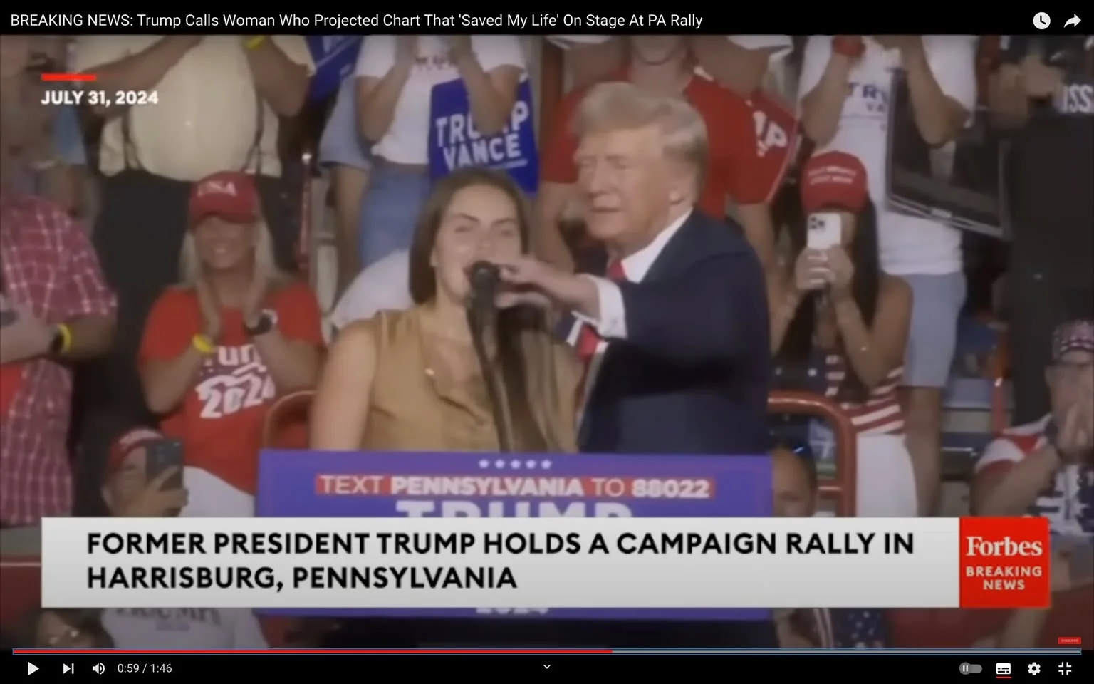 Former President Trump is visible on stage at a campaign rally in Harrisburg, Pennsylvania. The event takes place indoors, with a crowd of supporters in the background. Trump, wearing a dark suit with a red tie, appears to be introducing or speaking with a woman beside him. The woman is wearing a sleeveless brown top and is positioned at the podium where Trump is holding a microphone. The crowd is engaged, with some individuals wearing red hats and shirts displaying support for Trump. A large sign on the podium reads "TEXT PENNSYLVANIA TO 88022," and a banner below reports that Trump is holding a campaign rally. The date "JULY 31, 2024" is displayed prominently on the screen. The lighting is bright, typical of a public speaking event, and the atmosphere is energetic, with supporters clapping and taking photos. The video is from the "Forbes Breaking News" channel on YouTube, highlighting the significance of the rally as a notable political event.