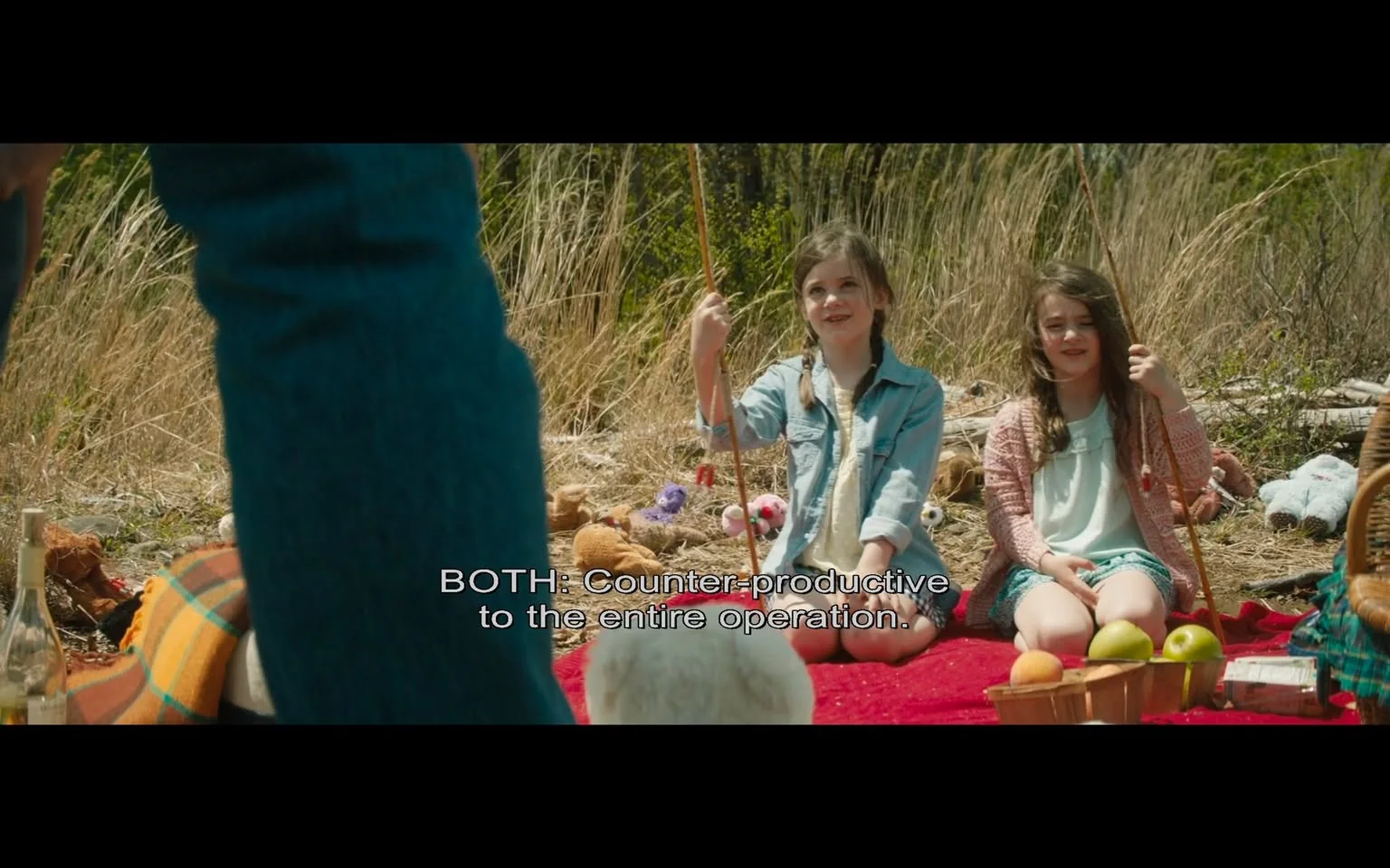 This video frame depicts an outdoor scene during the daytime, featuring two young girls seated on a red blanket. They appear to be having a picnic, surrounded by various items like stuffed animals, baskets, and fruit, including apples. The setting is a natural area with tall grasses in the background, suggesting a rural or park-like environment. The girls, sitting cross-legged, are holding sticks and seem to be engaged in a playful activity. One girl is wearing a denim jacket over a white dress, while the other is dressed in a light cardigan over a white dress. The overall atmosphere is casual and relaxed, with a mix of warm and natural colors. The foreground shows part of an adult's leg, indicating the presence of another person observing or interacting with the children. The caption in the image reads, "BOTH: Counter-productive to the entire operation," suggesting a conversation or narrative element related to the scene.
