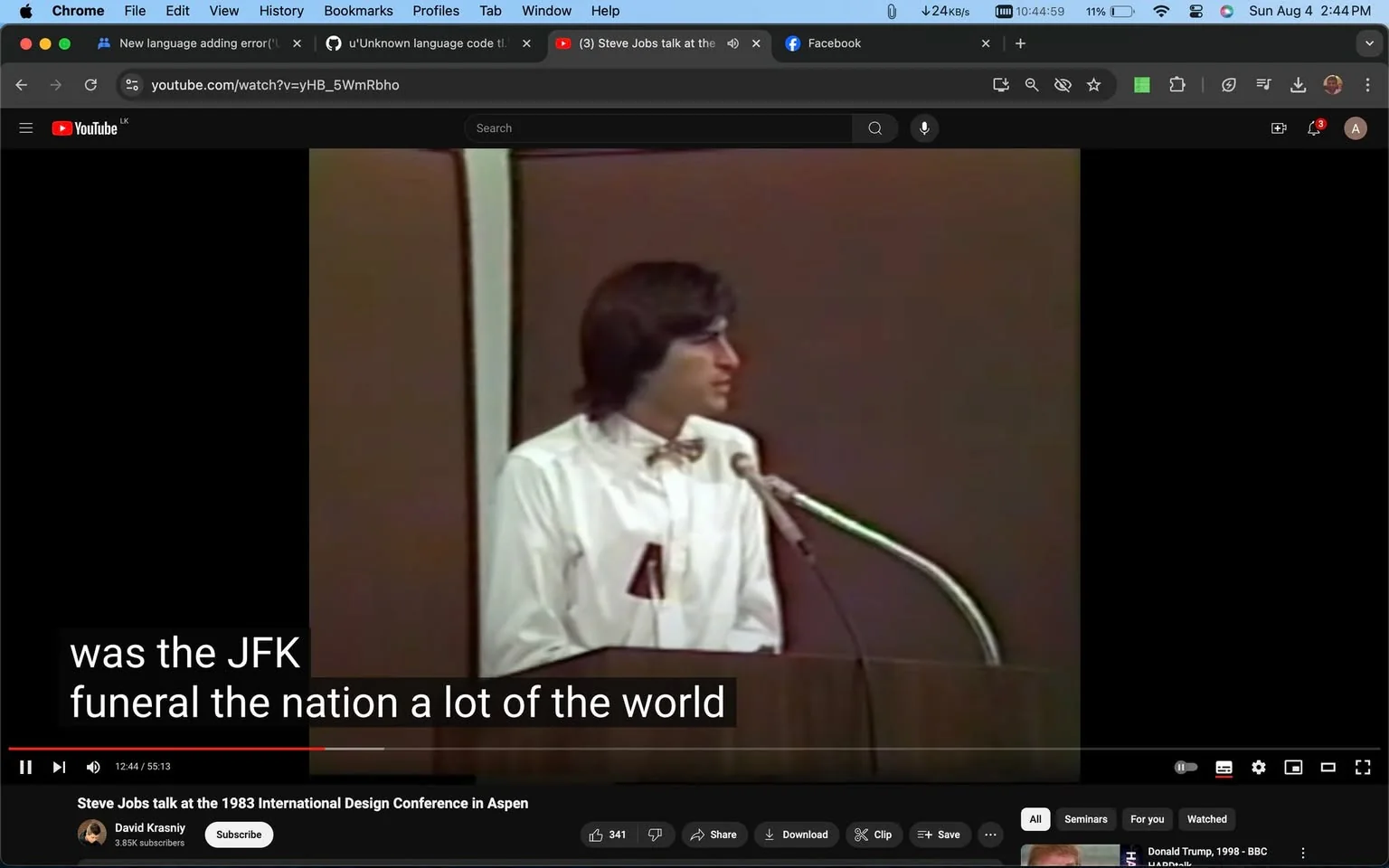 Steve Jobs is seen speaking at a podium during the 1983 International Design Conference in Aspen. The setting is indoor, with a plain brown backdrop behind him, suggesting a formal conference environment. Steve Jobs is wearing a white shirt with a bow tie, adding to the professional atmosphere. He is positioned slightly to the side, speaking into a microphone on a stand, indicating he is addressing an audience. The video is hosted on YouTube and is titled "Steve Jobs talk at the 1983 International Design Conference in Aspen," published by the channel David Krasniy. The on-screen text includes subtitles that read "was the JFK funeral the nation a lot of the world." This suggests that the content of the talk may be covering significant historical or cultural events. The overall tone appears informative and serious, typical of a conference presentation.