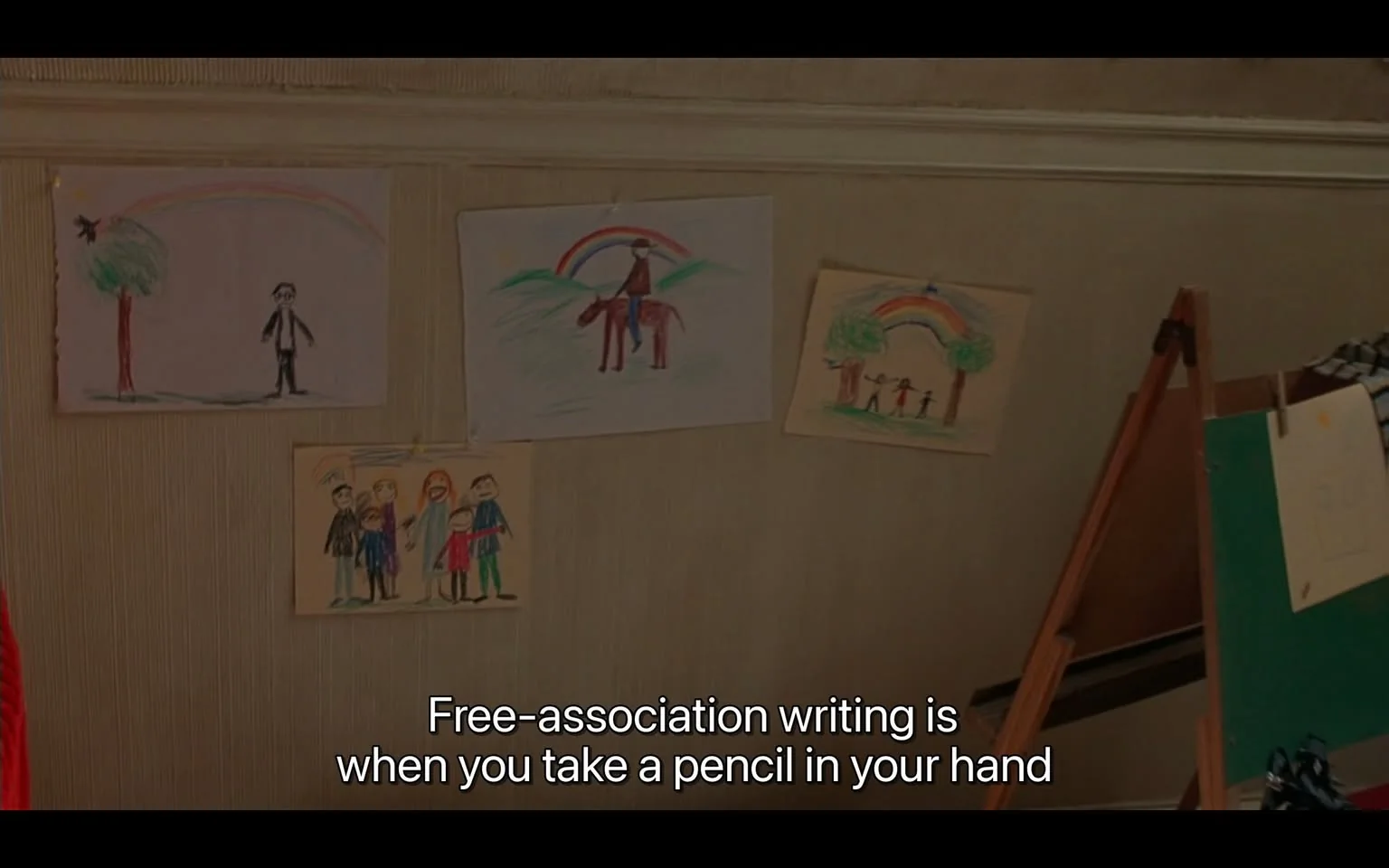 The image depicts a wall adorned with children's drawings, suggesting a warm and creative environment. Four distinct artworks are displayed, each featuring vibrant colors and simple figures. The drawings include scenes such as a person standing by a tree with a rainbow overhead, a figure on a horse beneath a rainbow, a group of people under a rainbow between two trees, and a family group portrait. The paper is tacked to the wall, indicating a casual display likely in a home or classroom setting. An easel is visible to the right, holding more paper, reinforcing the artistic theme.
The setting is likely a child's room or an art space. The lighting is soft, contributing to a cozy atmosphere. The captured text at the bottom reads, "Free-association writing is when you take a pencil in your hand," suggesting a moment of explanation or learning. This phrase complements the creative theme, indicating perhaps a lesson or discussion happening off-screen about artistic or expressive practices.