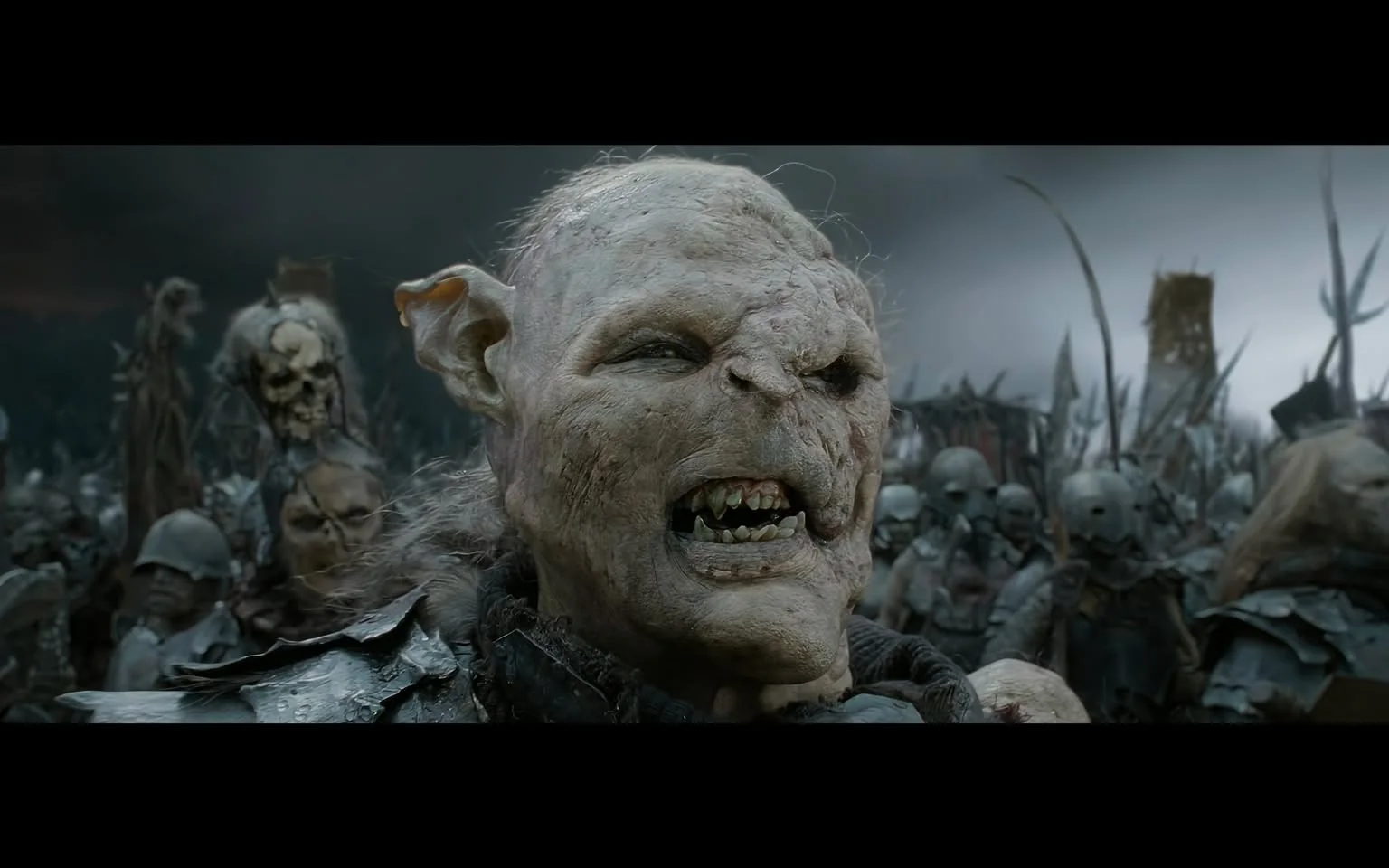 The image depicts a scene from "The Lord of the Rings," showcasing a fierce and intimidating orc from the franchise. The orc is prominently featured in the foreground, characterized by its grotesque and distorted facial features, including a misshapen nose and uneven teeth. Its skin is a pallid, rough texture, adding to its menacing appearance. The background is filled with a mass of other orcs and warriors, indicating a large army poised for battle. They are clad in dark, rough armor, holding spears and weapons, creating a sense of impending conflict. The lighting is dim and overcast, with a foreboding sky that emphasizes the grim atmosphere of the impending battle. The composition and style reflect the epic and dark fantasy elements typical of the film series, immersing the viewer in the chaotic and dangerous world of Middle-earth. This image captures the tension and scale of the battles central to the storyline.