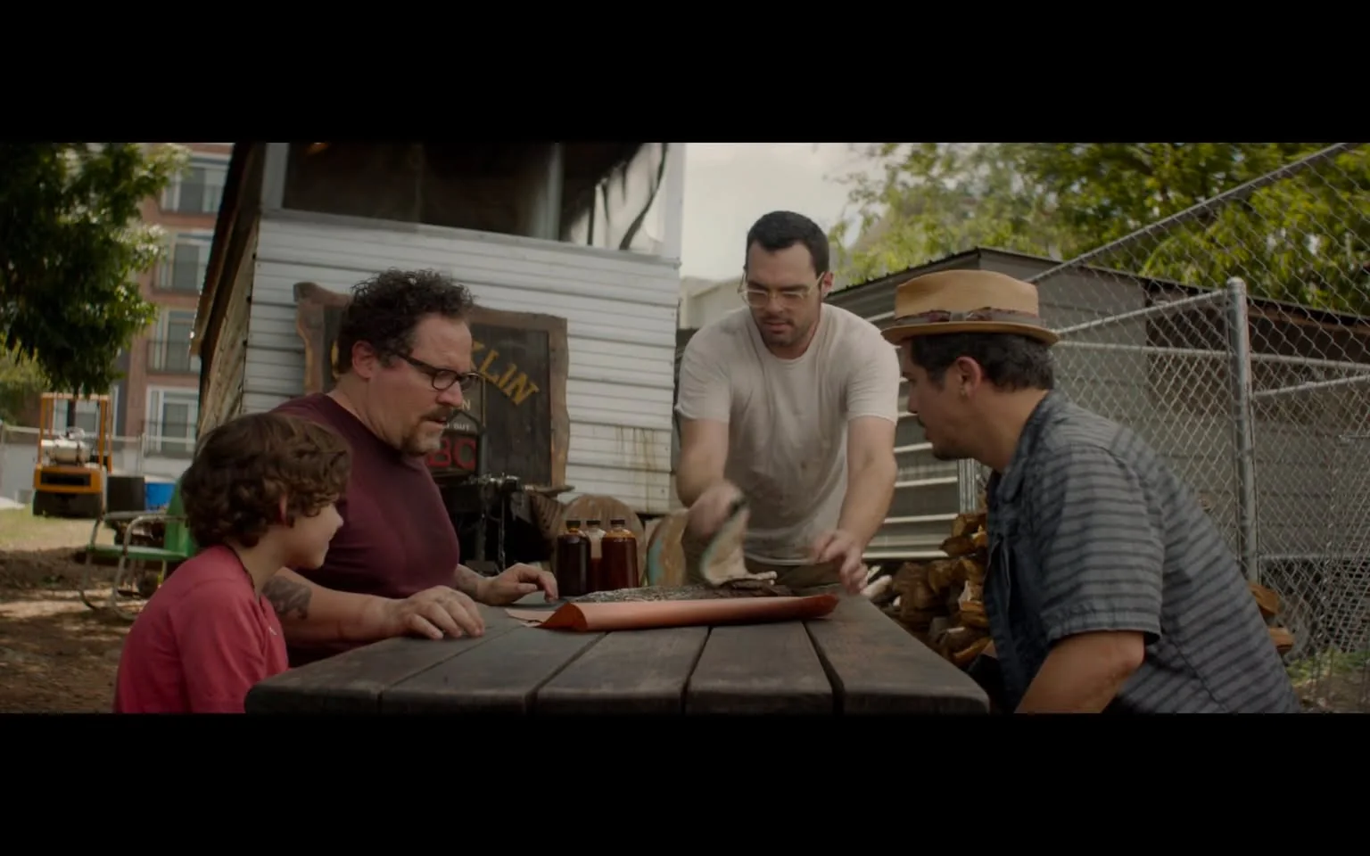 In this image, Aaron Franklin is prominently featured in the center, representing the best of Franklin BBQ. He is handing food to a group seated around a wooden picnic table situated outdoors. The setting is casual and relaxed, with trees and a building in the background, suggesting a friendly and communal atmosphere. Aaron, wearing a white t-shirt, appears engaged and hospitable as he serves a dish wrapped in butcher paper, typical of BBQ presentation.
To his right, a man in a striped shirt and straw hat leans forward, showing interest in the meal. On the left, two others are seated; one is a child in a red shirt and the other is a man with glasses and a beard, wearing a dark red t-shirt, both focused on the food. The table is adorned with BBQ sauce bottles and drinks, emphasizing a casual dining experience. The lighting is natural daylight, enhancing the warm and inviting mood of the scene. Behind them, a sign partially visible reads "Franklin," further cementing the BBQ theme.
- Key OCR Lines: "FRANKLIN"