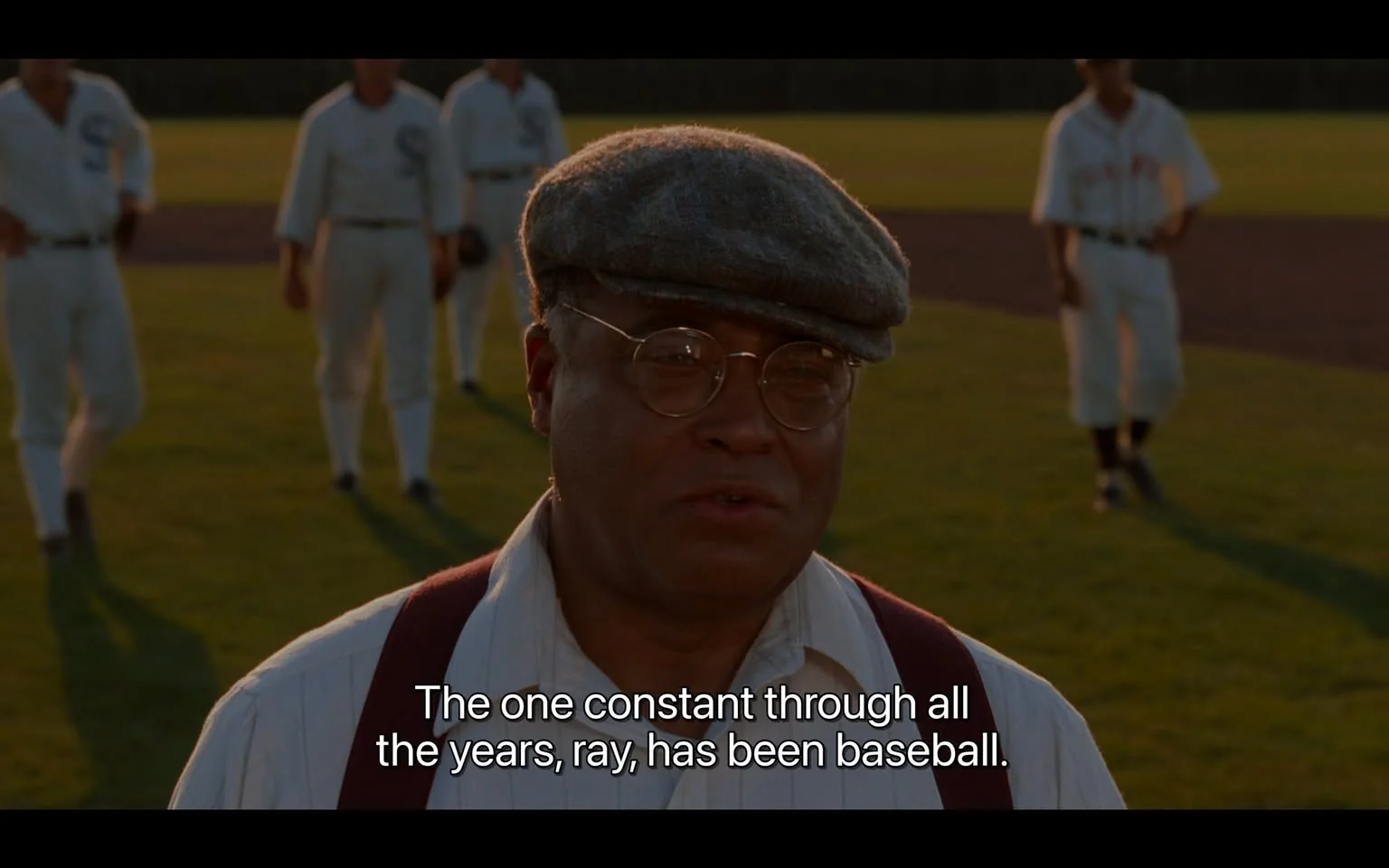 The image features a scene from the film "Field of Dreams" with James Earl Jones in the foreground. He is wearing a flat cap, glasses, and a white shirt with dark suspenders. His expression appears thoughtful and earnest as he speaks. Behind him, several men in vintage baseball uniforms are visible, suggesting a historical or nostalgic setting related to baseball. The background is a lush green field, which enhances the sense of an outdoor, daytime scene. The lighting is warm, casting soft shadows on the characters, emphasizing a serene and reflective atmosphere. The caption reads, "The one constant through all the years, ray, has been baseball," highlighting a key theme of the film related to the timeless nature of the sport. James Earl Jones's character is central to conveying this idea, adding depth to the narrative with his presence and delivery.
- Captions / subtitles: "The one constant through all the years, ray, has been baseball."