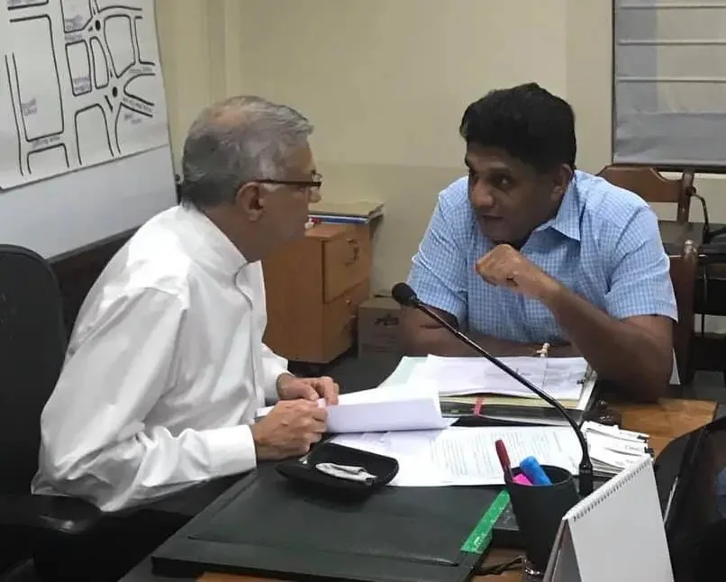 Ranil Wickremasinghe and Sajith Premadasa are engaged in a conversation in an office setting. Ranil Wickremasinghe, wearing a white shirt, is seated on the left side of the image. He is facing Sajith Premadasa, who is on the right, wearing a blue checkered shirt. They are both leaning towards each other, indicating a serious discussion. Several documents and papers are spread out on the table between them, suggesting the meeting's focus on paperwork or official matters. A microphone is positioned between them, possibly for recording or amplifying the conversation. The room contains office furniture like desks and chairs, and a map or chart is partially visible on the wall behind them. The atmosphere appears professional and focused, with both individuals seemingly engaged in the topic at hand. The lighting is neutral, typical of an indoor office environment.