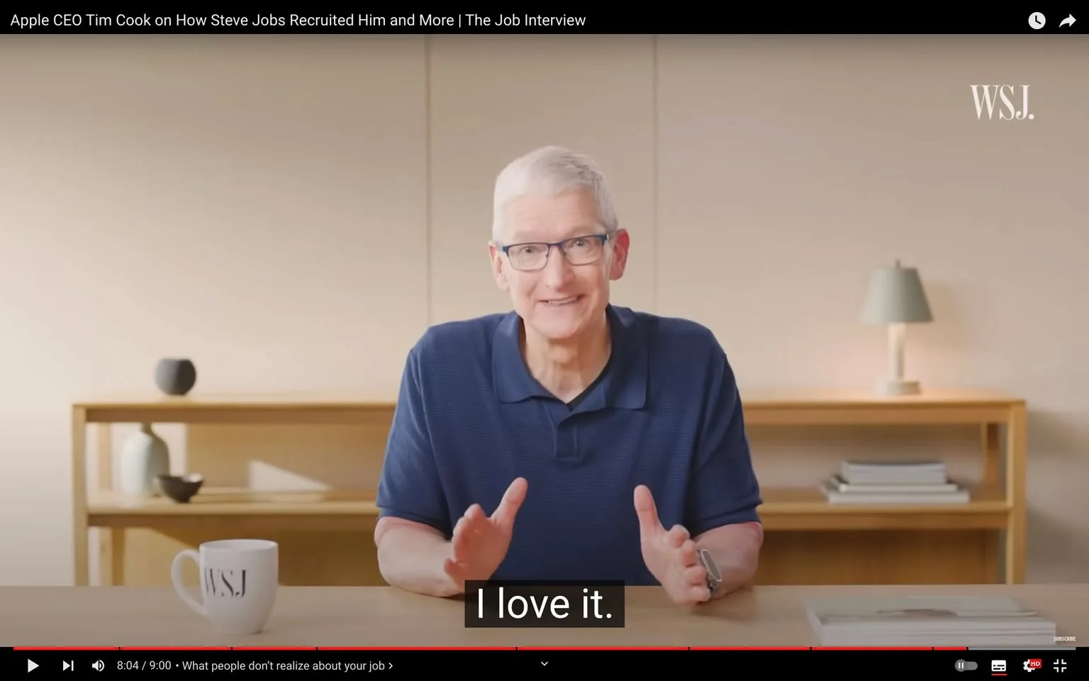 Tim Cook is featured in this video frame from an interview titled "Apple CEO Tim Cook on How Steve Jobs Recruited Him and More | The Job Interview," available on YouTube. He is sitting at a simple wooden table in a minimalist setting, likely an office or studio. Tim Cook is wearing a navy blue polo shirt, and his demeanor is relaxed and approachable. His hands are slightly raised as he gestures during the conversation. The room has a soft, neutral color palette, with a few decorative items such as a small vase and a lamp on a shelf behind him. A mug with the "WSJ" logo is placed on the table, suggesting the interview is conducted by the Wall Street Journal. The lighting is bright and evenly distributed, enhancing the professional yet casual atmosphere. Subtitles appear at the bottom, with the text "I love it," indicating a moment of enthusiasm or appreciation in his dialogue.
- Title: "Apple CEO Tim Cook on How Steve Jobs Recruited Him and More | The Job Interview"
- Channel / profile: "WSJ"
- Site / app: "YouTube"
- Captions / subtitles: "I love it."