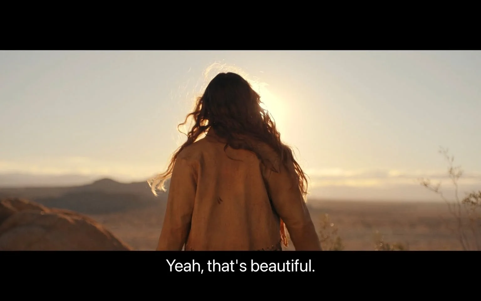 The image shows a person with long, wavy hair standing in a desert landscape during sunset. The sun is positioned directly behind them, creating a striking halo effect around their head. They are wearing a brown jacket, and the lighting casts a warm glow on the scene, emphasizing the serene and contemplative mood. The vast expanse of the desert stretches out in the background, with distant hills and a clear sky, adding to the sense of openness and tranquility. The composition highlights the natural beauty of the setting, aligning with the subtitle: "Yeah, that's beautiful." This scene captures a moment of quiet reflection amidst the backdrop of a stunning sunset.