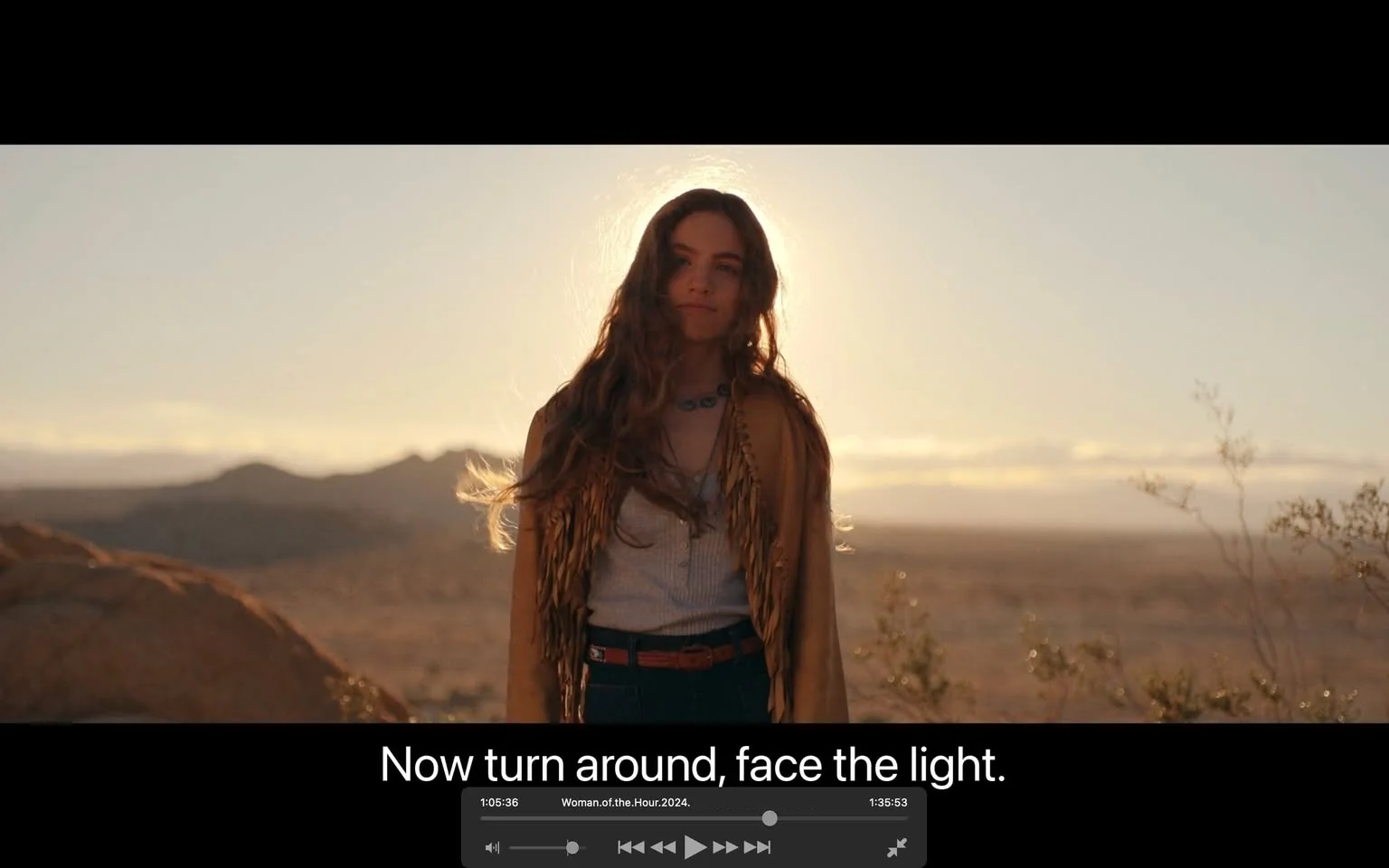 The video frame is from the film "Woman of the Hour" and features an outdoor scene set during sunset in a desert landscape. A young woman is positioned in the foreground, standing against a backdrop of rolling hills and a wide, open sky. She has long, wavy hair and is wearing a fringed jacket over a light-colored top, paired with jeans. A necklace adorns her neck, adding a touch of bohemian flair to her appearance. The lighting is warm and soft, with the setting sun creating a halo effect around her head, emphasizing the serene and contemplative mood of the scene. The subtitle at the bottom reads, "Now turn around, face the light," suggesting a moment of reflection or revelation. The composition captures a tranquil and introspective atmosphere, with the vastness of the desert emphasizing the character's solitude or journey.
- Captions / subtitles: "Now turn around, face the light."