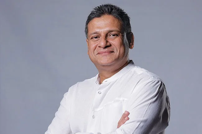 Dilith Jayaweera is depicted in this professional studio portrait. He is wearing a crisp white shirt with a standing collar, which adds to his polished and formal appearance. The background is a simple, neutral gray, ensuring the focus remains on the subject. Dilith Jayaweera is standing with his arms crossed, projecting confidence and approachability. His expression is warm and composed, with a slight smile that suggests openness and friendliness. The lighting is soft and even, illuminating his face without harsh shadows, enhancing the clarity and detail of the portrait. This image likely serves a business or professional purpose, reflecting a sense of authority and competence.