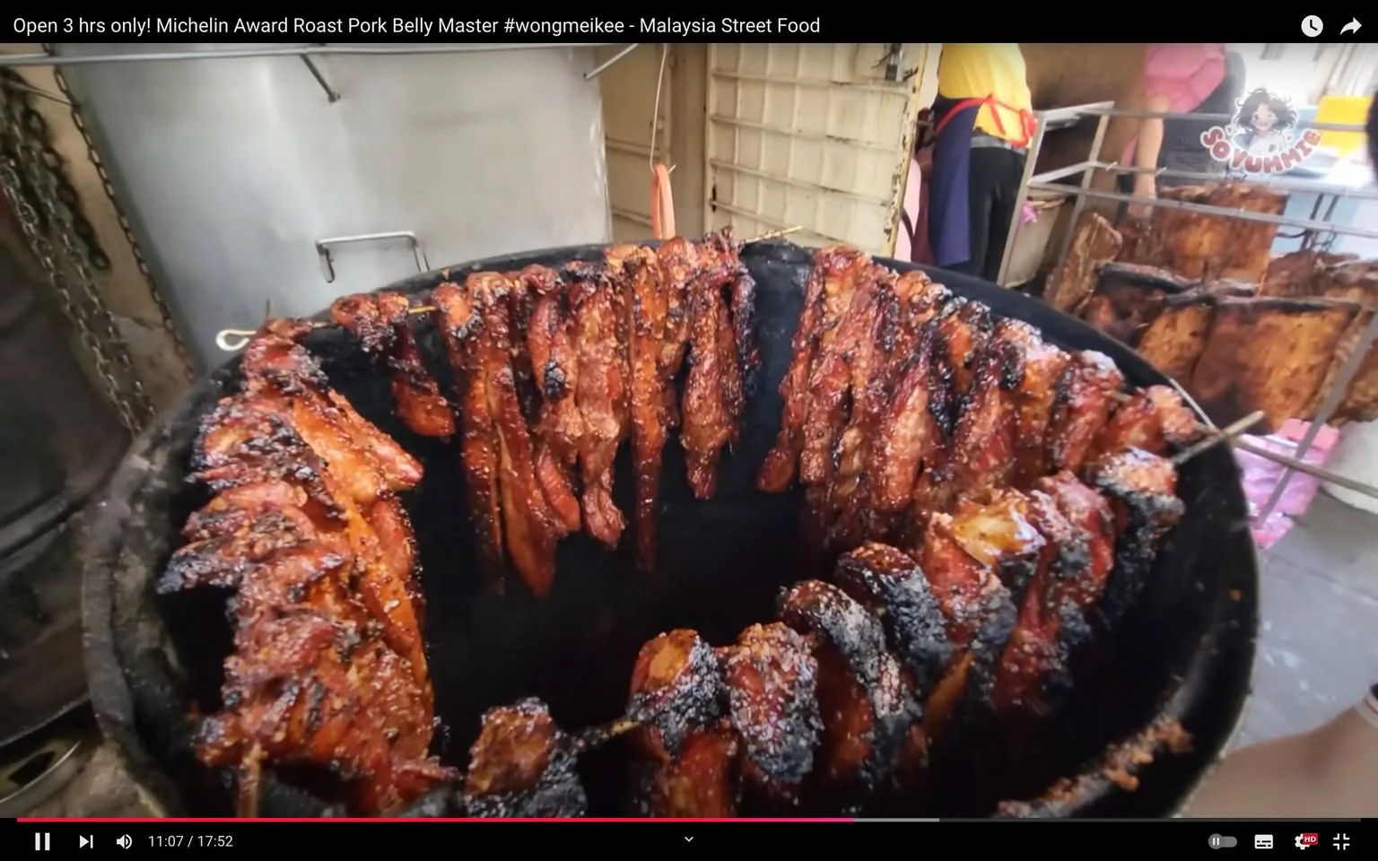 The image is a video frame showcasing a bustling street food scene, highlighting the preparation of roast pork belly. The setting is outdoors, likely at a market or a street food stall in Malaysia. In the center of the frame, numerous skewers of pork belly are hung vertically around the interior of a large roasting drum, each piece glistening with a caramelized, golden-brown glaze, indicating they are being roasted to perfection. The texture of the pork suggests it is both juicy and crispy, with charred edges that promise a smoky flavor.
In the background, there are glimpses of the bustling activity behind the cooking area, where a person is partially visible, wearing a yellow shirt and a red apron, engaged in food preparation tasks. The environment is lively and informal, characteristic of a street food setting, with the presence of metal hooks and chains supporting the cooking apparatus. The lighting is natural, enhancing the rich colors of the roasted pork against the darker tones of the roasting drum.
The video is titled "Open 3 hrs only! Michelin Award Roast Pork Belly Master #wongmeikee - Malaysia Street Food," suggesting the featured food item has received acclaim, perhaps from a Michelin-awarded vendor. The frame is from a video associated with the channel "So Yummy," which likely focuses on diverse and delectable culinary experiences. The scene captures the essence of street food culture, where the art of roasting is both a visual and sensory delight.