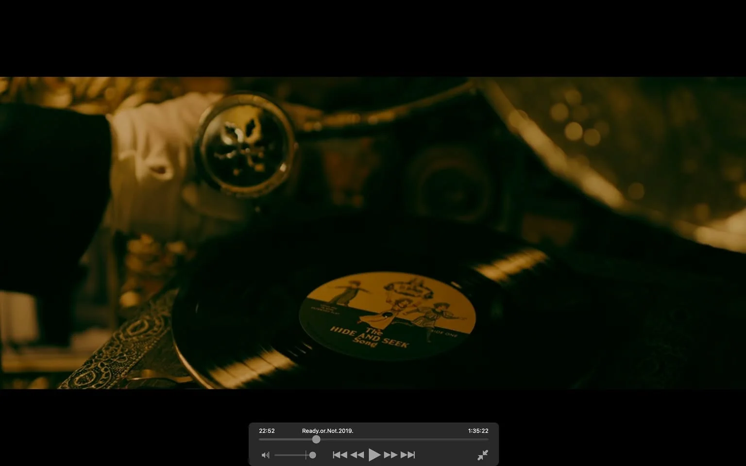 The image depicts a close-up of a vinyl record being placed on a turntable. The scene is from the movie "Ready or Not," as indicated by the file name and timestamp. The lighting is dim, casting a warm, vintage hue over the scene, enhancing the antique feel of the setting. A gloved hand can be seen adjusting the tonearm, which has an ornate, metallic design featuring a skull. The vinyl's label is visible, displaying the title "The Hide and Seek Song" along with illustrations of people in playful poses, reminiscent of a bygone era. The setting appears to be indoors, with an evident focus on the nostalgic theme conveyed through the use of a traditional record player and the aged appearance of the surroundings. The intricate details of the turntable and the vintage aesthetic contribute to the eerie mood of the scene.