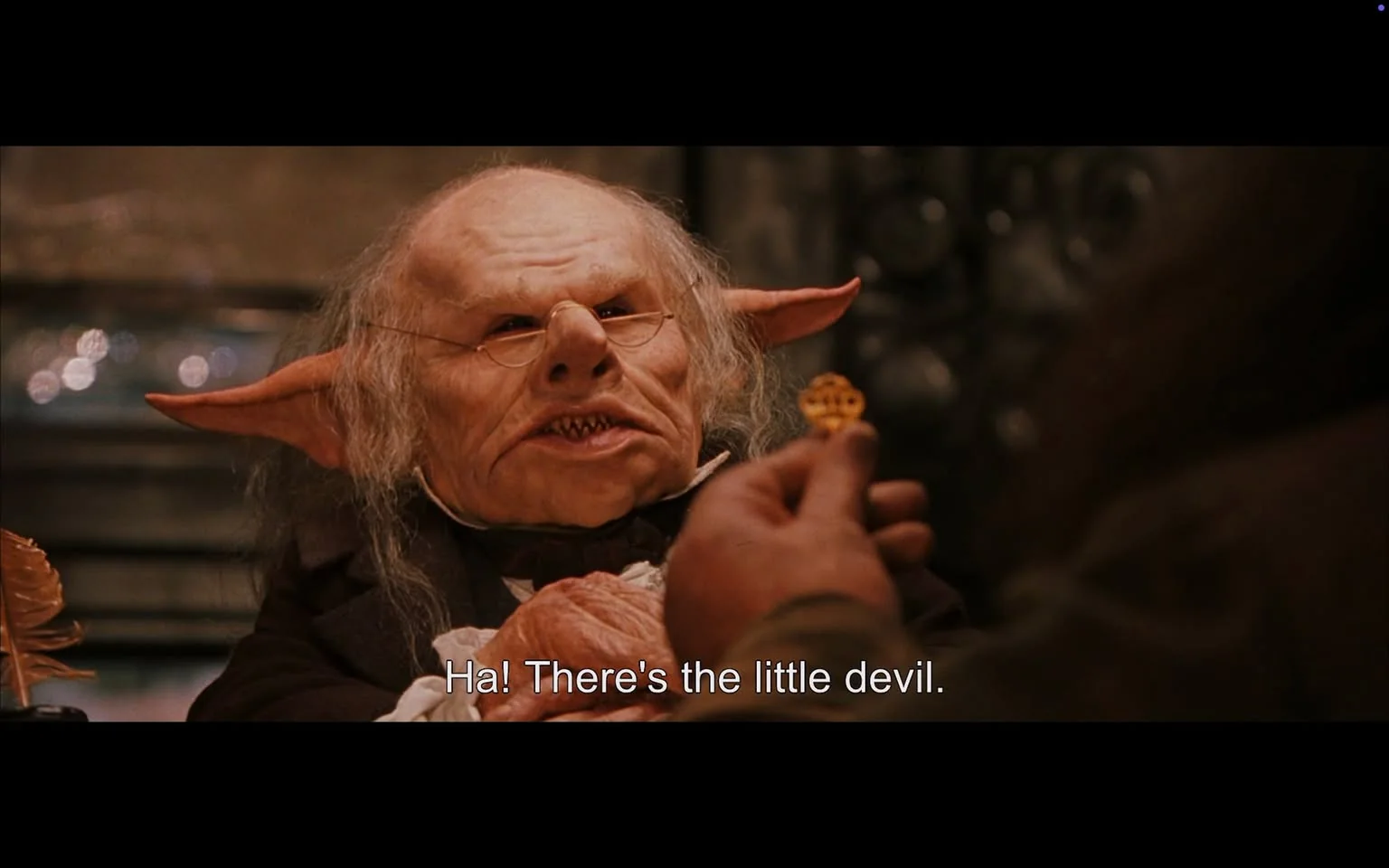 The image is a scene from the "Harry Potter" series, set in a fantasy environment. It shows a goblin-like creature with distinctive features, including large, pointed ears and a wrinkled face, wearing small glasses and a formal outfit. The creature is seated, looking at someone holding a small ornate key. The atmosphere appears to be a magical setting, likely in a wizarding bank, given the creature's attire and the key. The lighting is warm, highlighting the intricate details of the creature's face and clothing. The background is slightly blurred, focusing attention on the creature and the key in the hand. The scene carries a slightly mysterious and whimsical tone, characteristic of the magical world in "Harry Potter."

- Captions / subtitles: "Ha! There's the little devil."