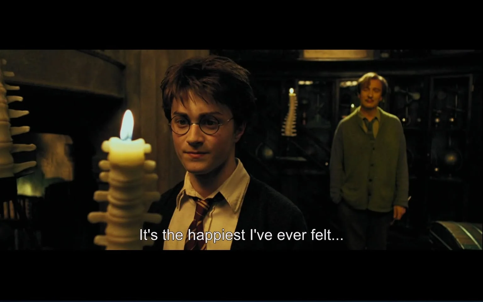 In this scene from a Harry Potter film, Harry Potter is prominently featured in the foreground, holding a uniquely layered candle that is lit, creating a warm glow that illuminates his face. He is wearing his Hogwarts uniform, which includes a white shirt and a maroon and gold striped tie, indicative of his Gryffindor house. His expression is one of contentment, matching the subtitle, "It's the happiest I've ever felt...," suggesting a moment of personal fulfillment or joy.

In the background, a man can be seen standing, dressed in a green cardigan over a shirt and tie, observing Harry with a gentle demeanor. The setting appears to be an indoor, dimly lit room, possibly a cozy, private study or classroom filled with mysterious objects and shelves, enhancing the magical atmosphere typical of the Harry Potter series. The lighting, dominated by the candle, creates a soft ambiance, emphasizing the intimacy and warmth of the scene.