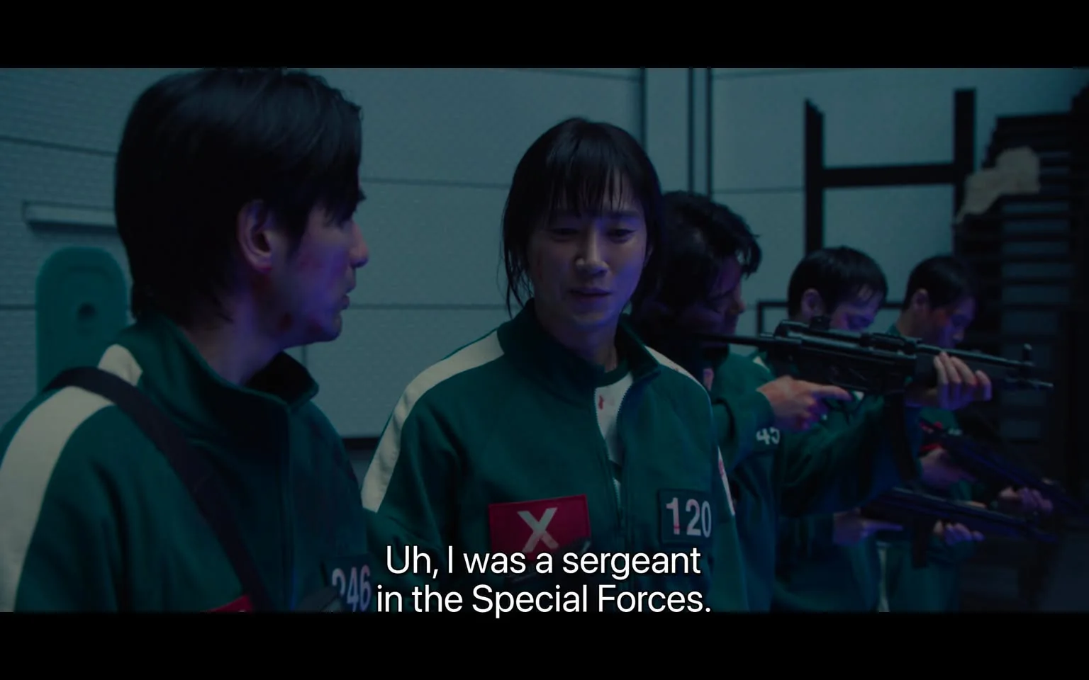 The image is a screenshot from the TV show "Squid Game." It features a dimly lit, indoor setting with characters wearing green tracksuits, which are iconic to the series. Each suit has a unique number visible on the front, indicating the players' identities within the game. The scene shows a group of people, some holding firearms, engaged in a conversation or preparing for a task. The individual in focus is speaking, as indicated by the subtitles: "Uh, I was a sergeant in the Special Forces." This line hints at the character's background and possibly their role in the scene. The lighting is cool and subdued, creating a tense and dramatic atmosphere typical of the show. The composition emphasizes the interaction between the characters, with attention to their expressions and body language, reflecting a moment of planning or strategy.