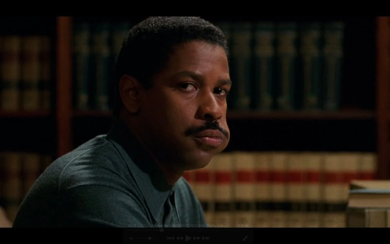 The image features a man seated at a table, facing slightly to the right. He has a medium complexion and short, curly black hair. His facial expression appears serious, with a slight frown and pursed lips. He has a prominent mustache that is well-groomed. The man is wearing a dark green, collared shirt, which is buttoned up. 

In the background, there are shelves filled with books, predominantly in dark colors, suggesting a library or office setting. The books have a uniform appearance, with some visible spines that appear to be leather-bound, possibly indicating they are law books or reference materials, given the context of the setting. 

The table in front of him is bare except for a small stack of papers or a book on the right side, and there is a slight reflection on the surface, indicating it may be made of a polished material. The lighting in the room is soft, creating a warm ambiance, and there are no visible windows or external light sources in the frame.

Overall, the setting conveys a professional atmosphere, likely related to legal or academic work, given the presence of books and the man's attire. There are no logos, trademarks, or readable text present in the image.